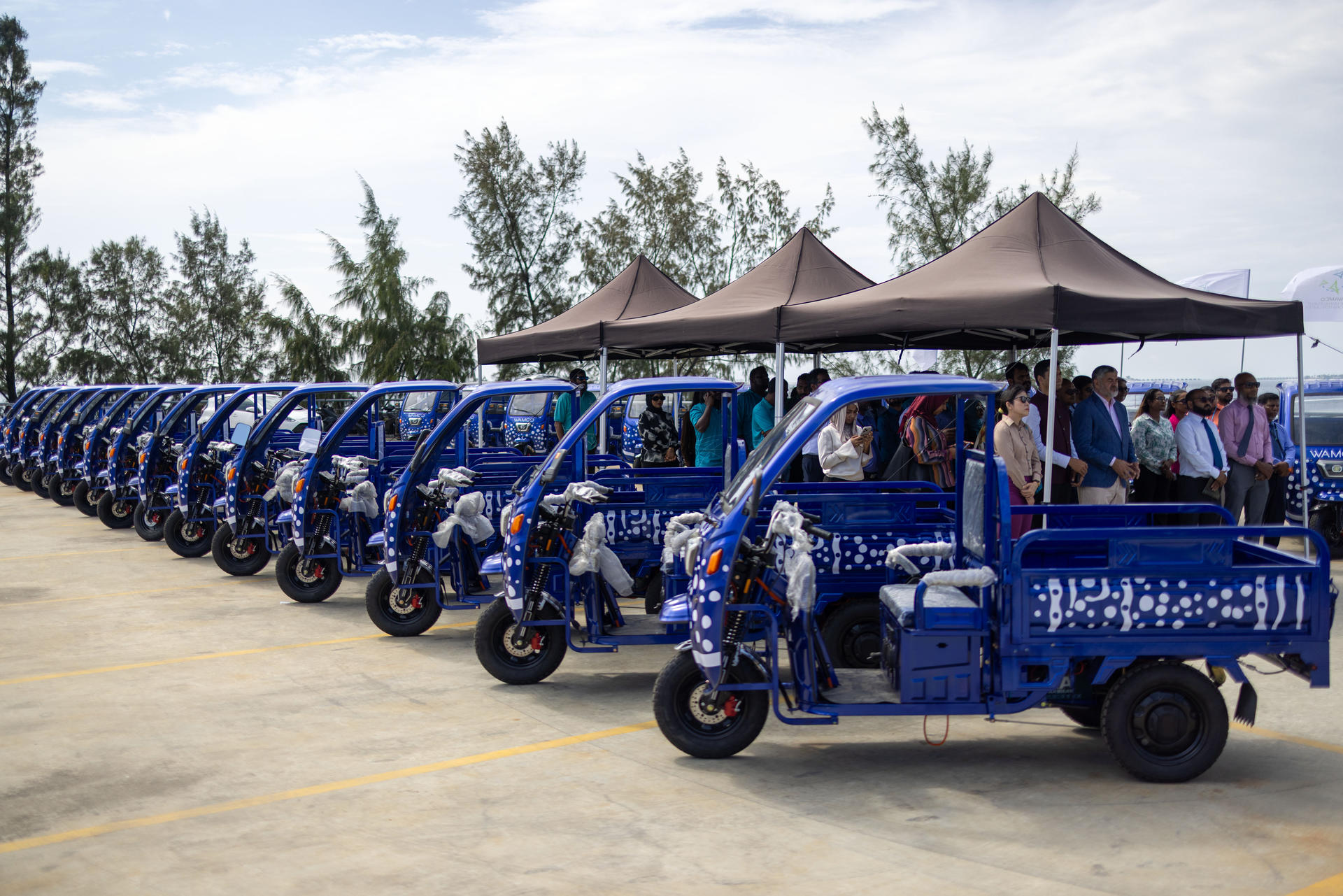 Row of blue e-vehicles (3 wheelers) parked under tents at an outdoor event.