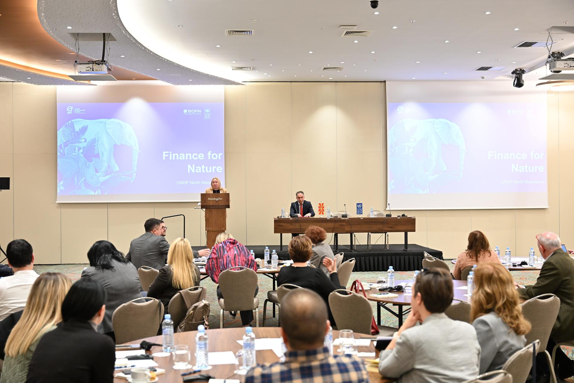 Conference room seminar with speaker at podium, panelists at a table, audience facing screens.