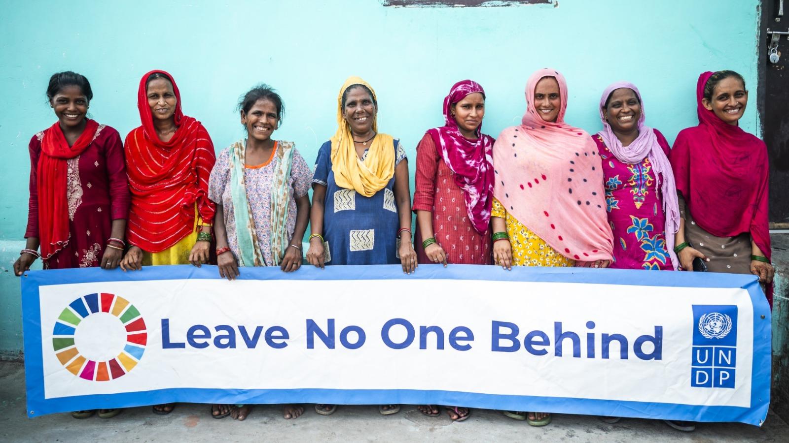 Group of people in colorful clothing standing behind a "Leave No One Behind" banner with SDG logo.