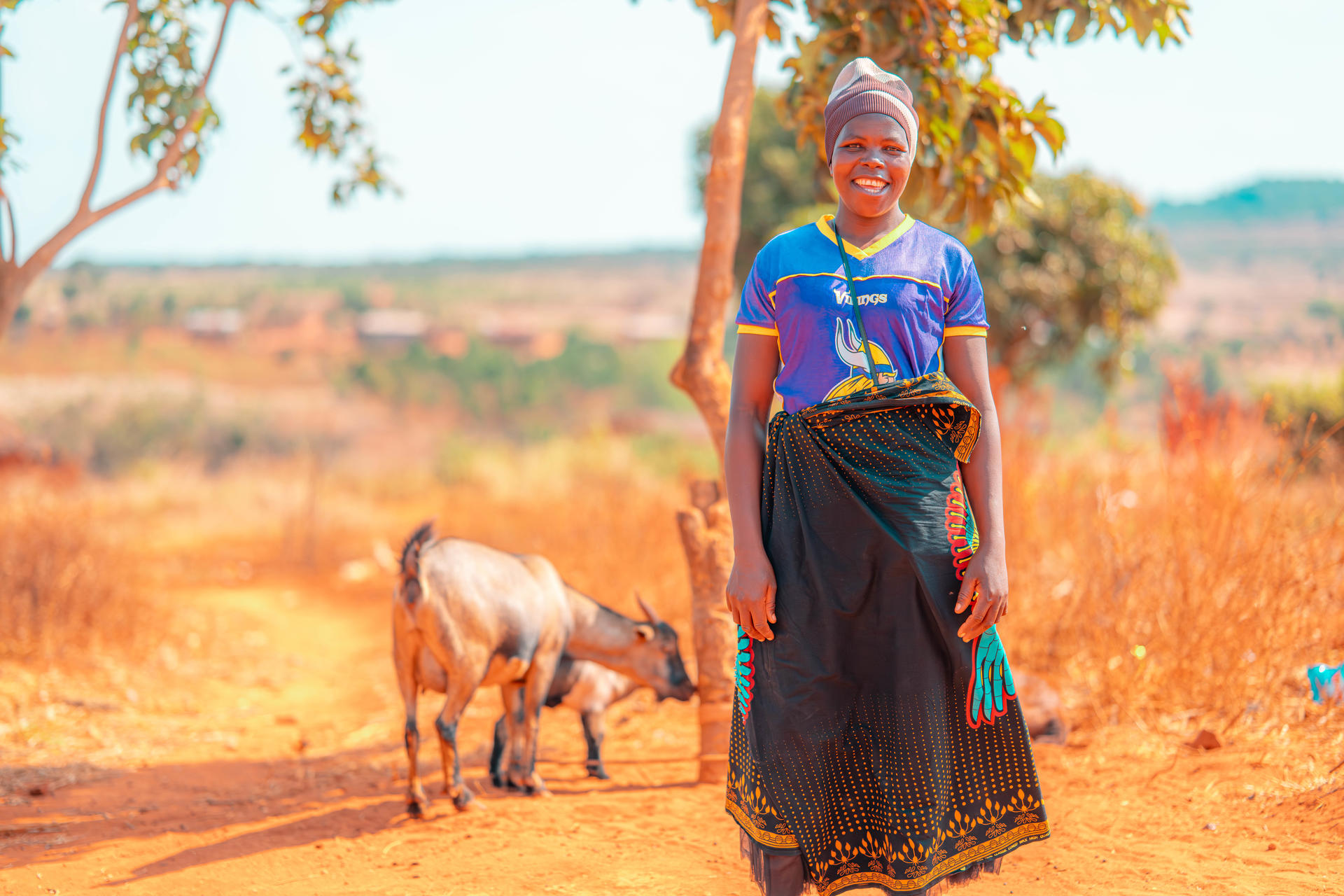 Person in a blue shirt and patterned skirt stands beside a small donkey in a dry rural landscape.