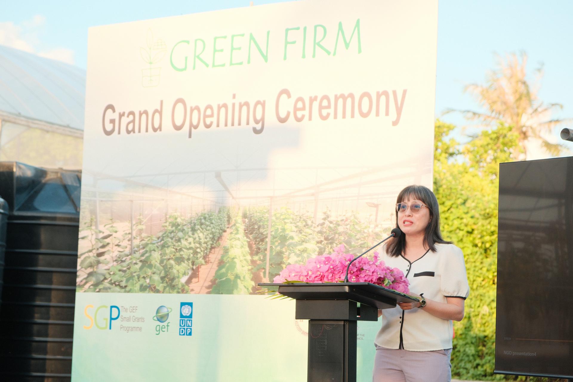 A woman at a podium delivering a speech at an outdoor grand opening; banner reads GREEN FIRM.