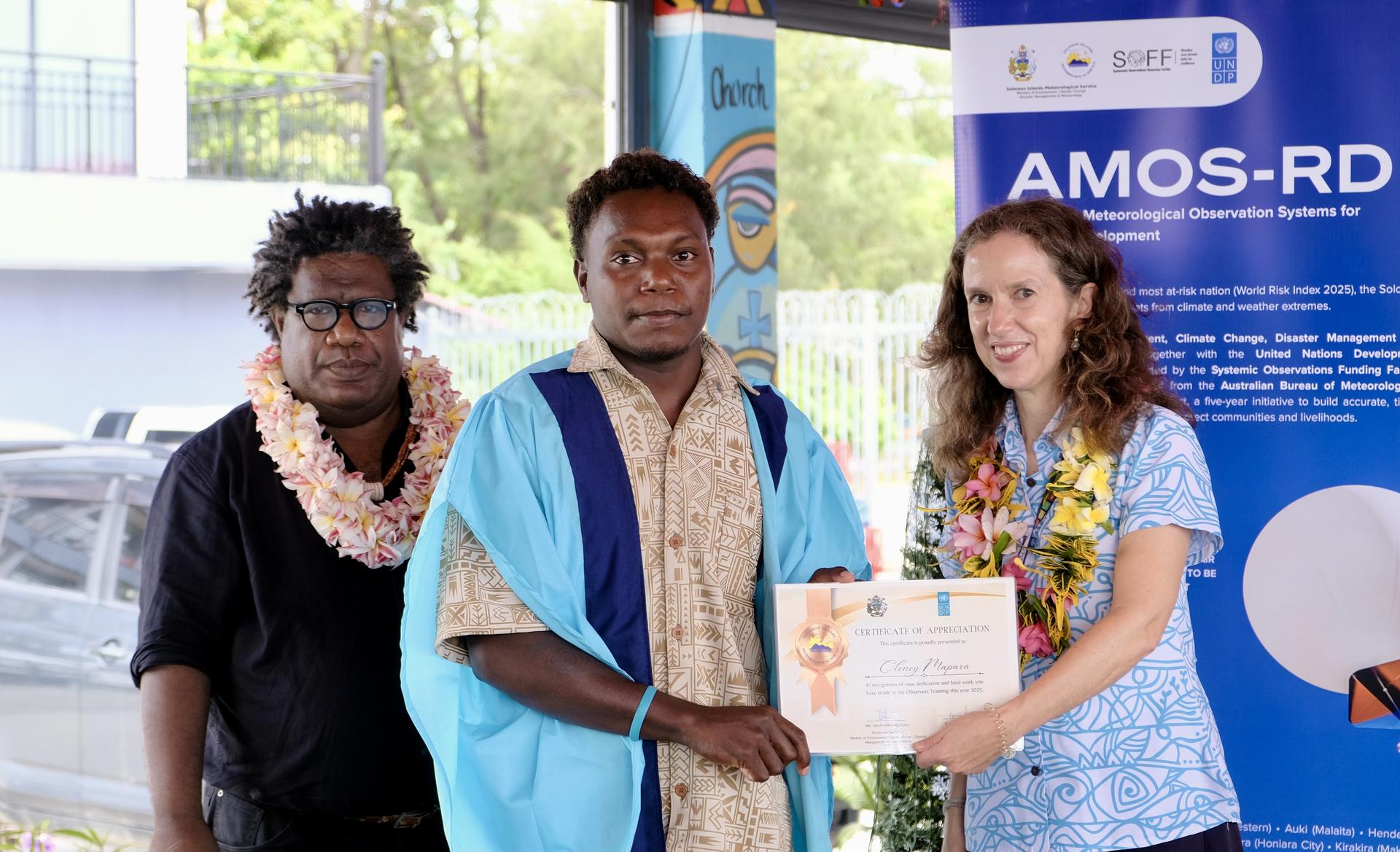 Graduate in blue gown receives certificate from a floral-dress presenter; blue banner behind.