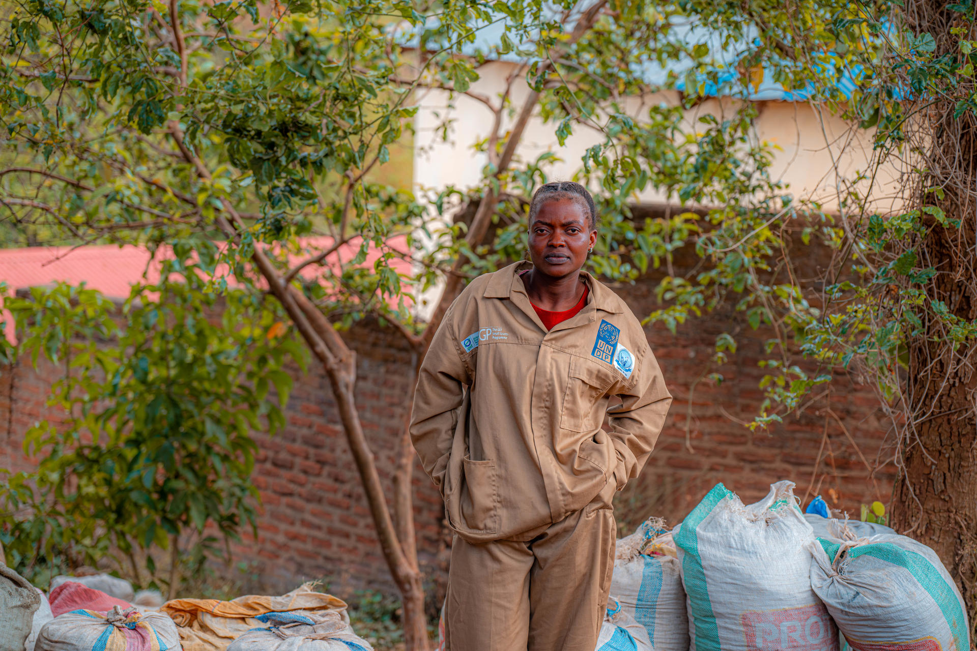 Man in tan coveralls stands outdoors beside bags near a vine-covered wall.
