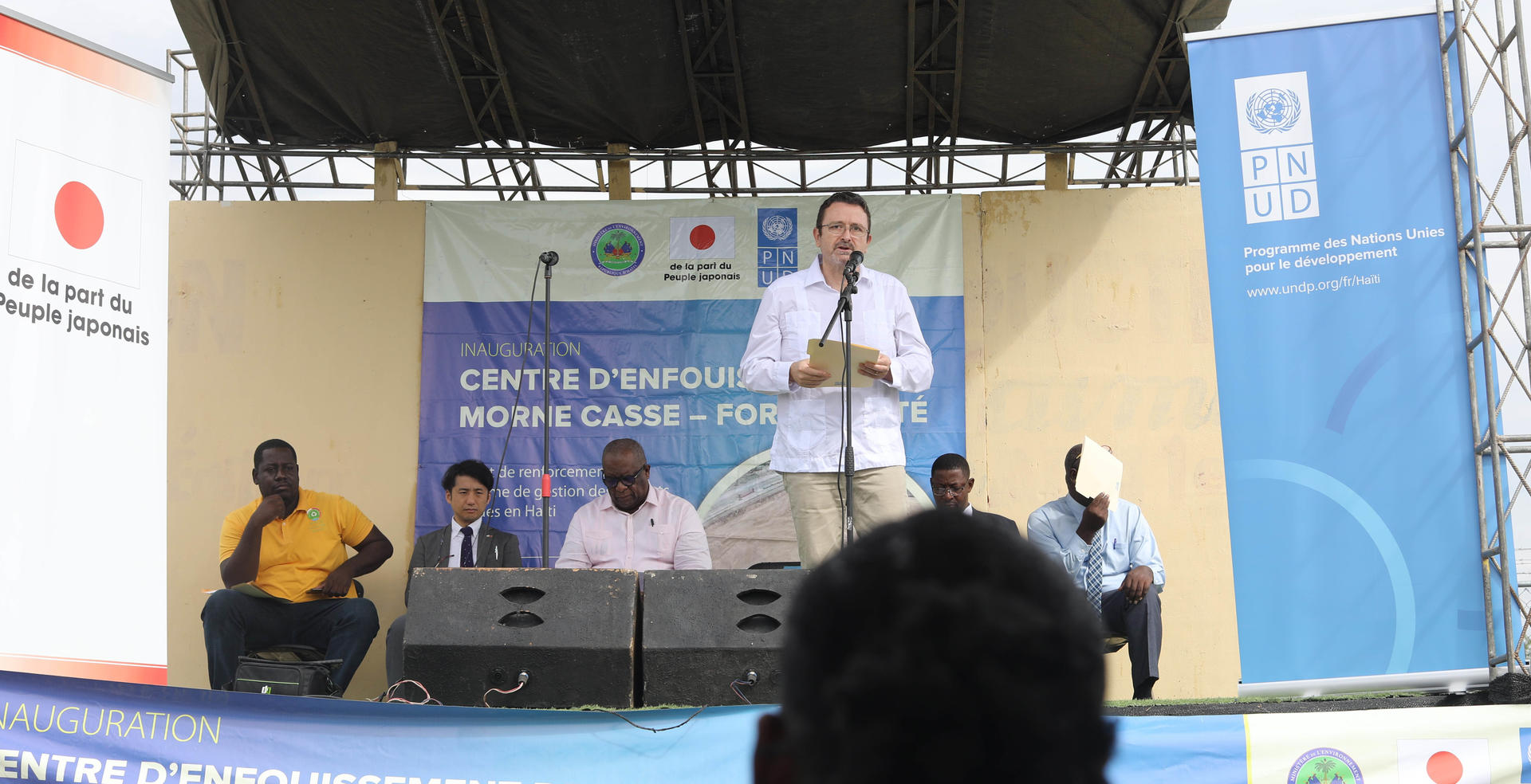 Photograph of a man speaking at a podium on an outdoor stage, panelists seated behind him, banners in view.