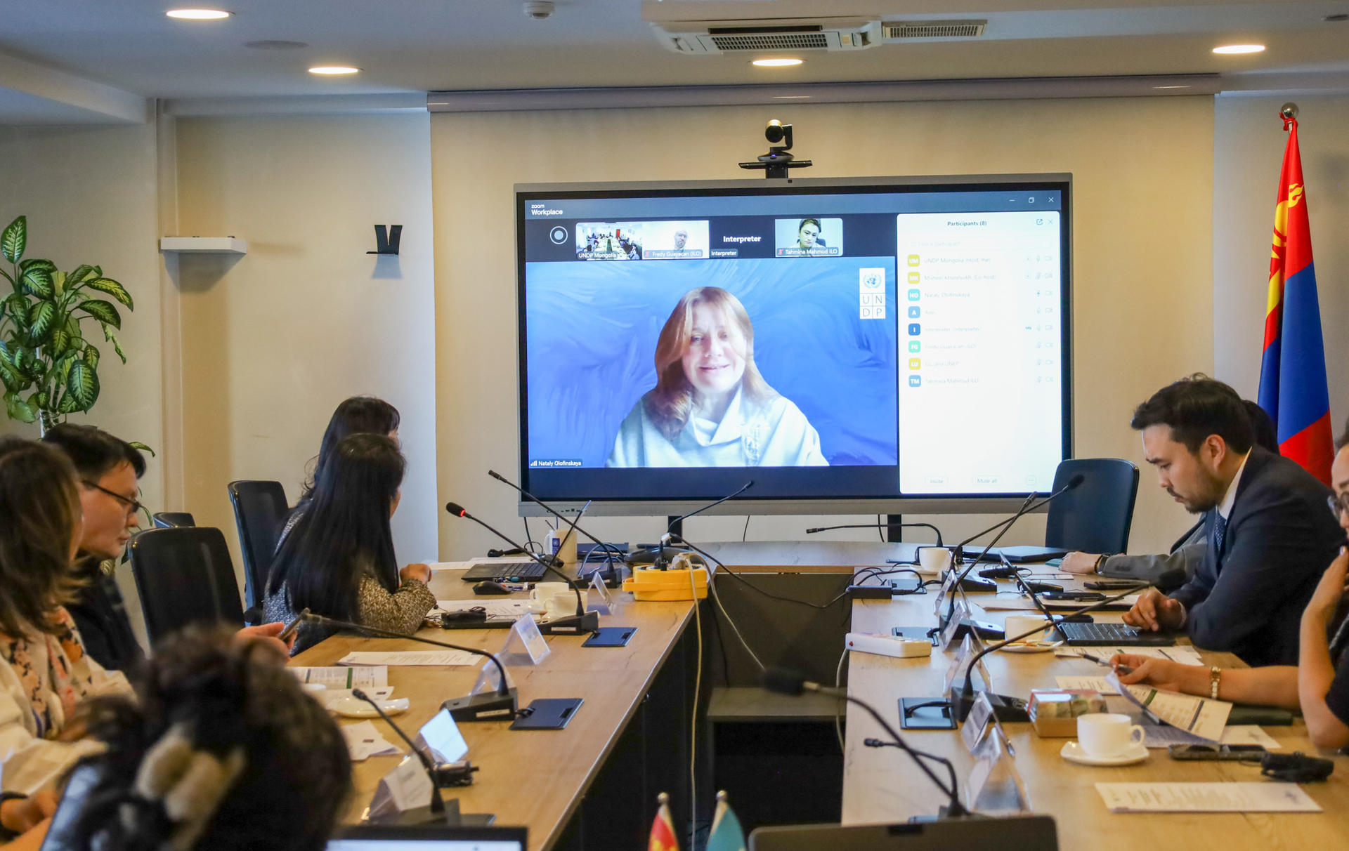 Conference room meeting; attendees around a long table; woman on video call speaks; Armenian flag.