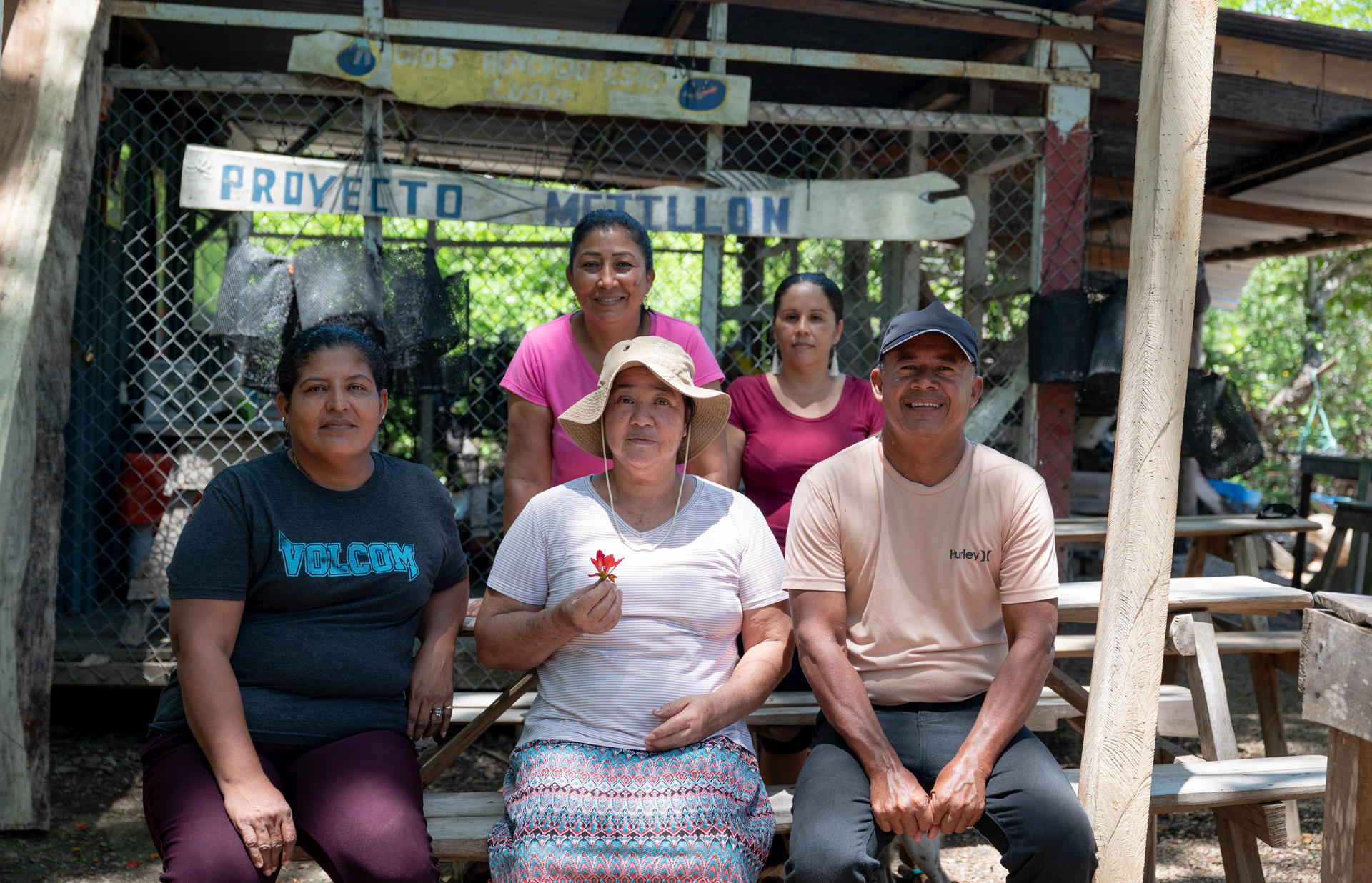 Five people sitting on a wooden bench under a shelter, smiling at the camera.