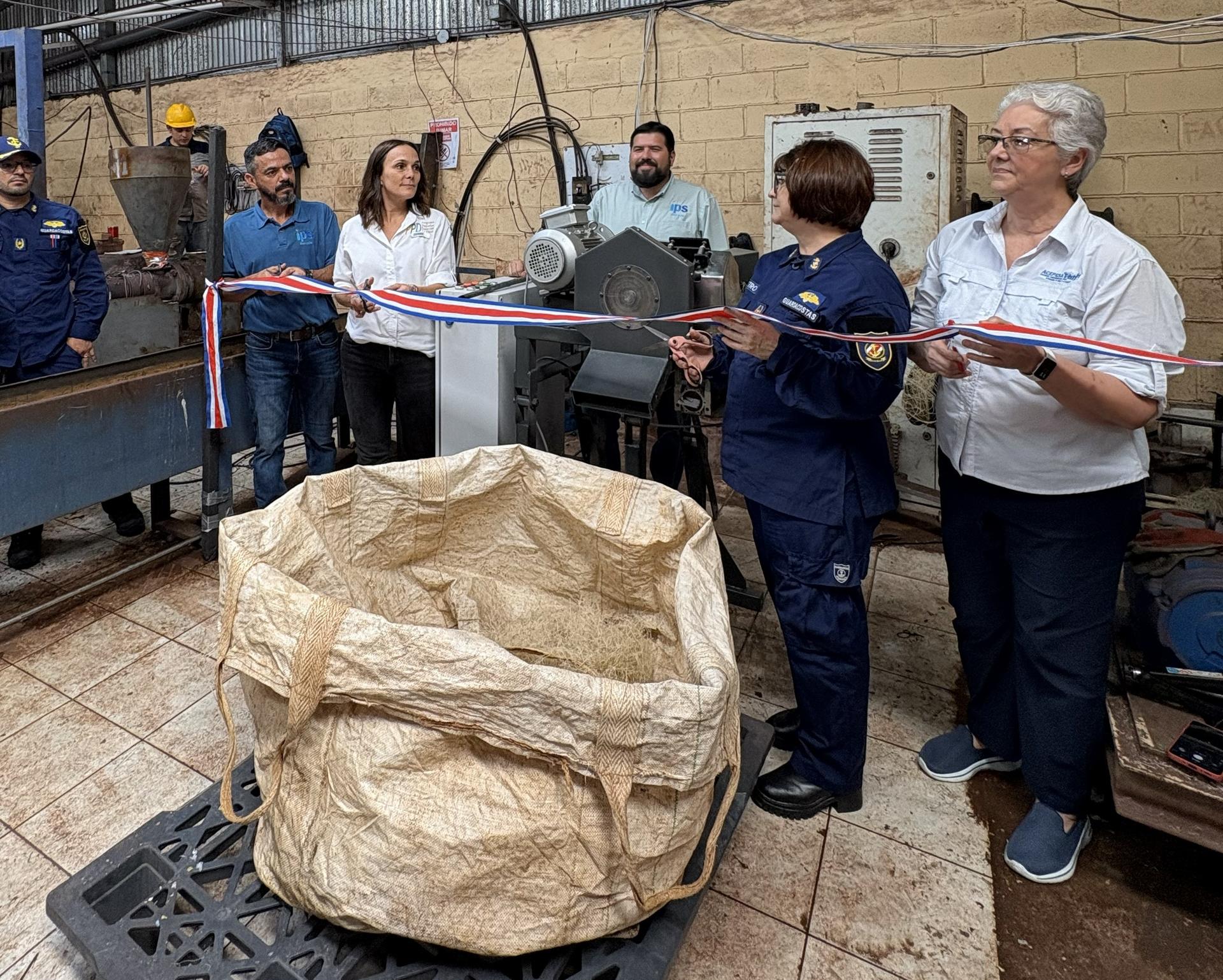 People in a workshop cut a red-and-white ribbon during a ceremony; brown paper bag on table.