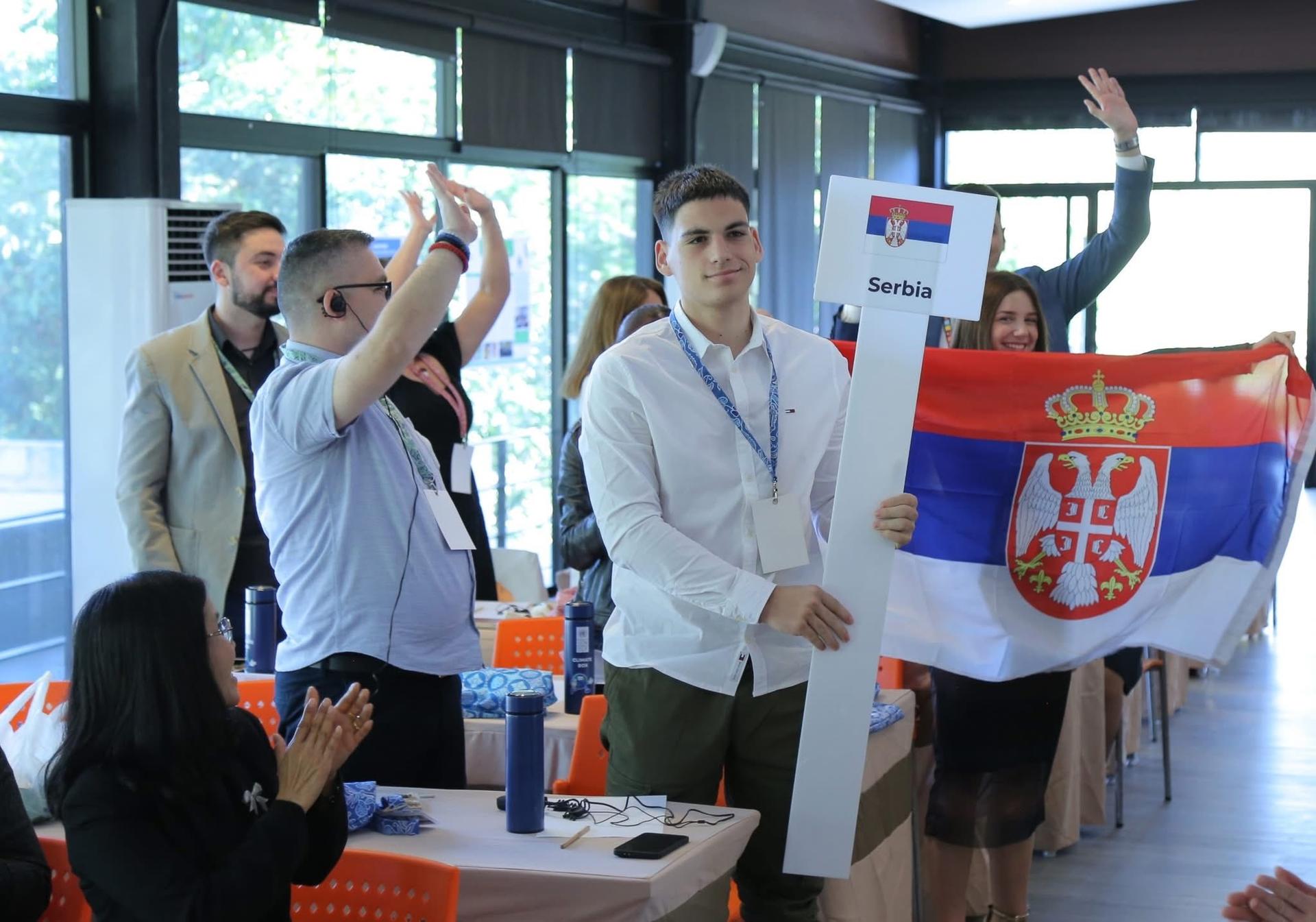 Group of people at an indoor event; man center raises hand; Serbian flag in background.