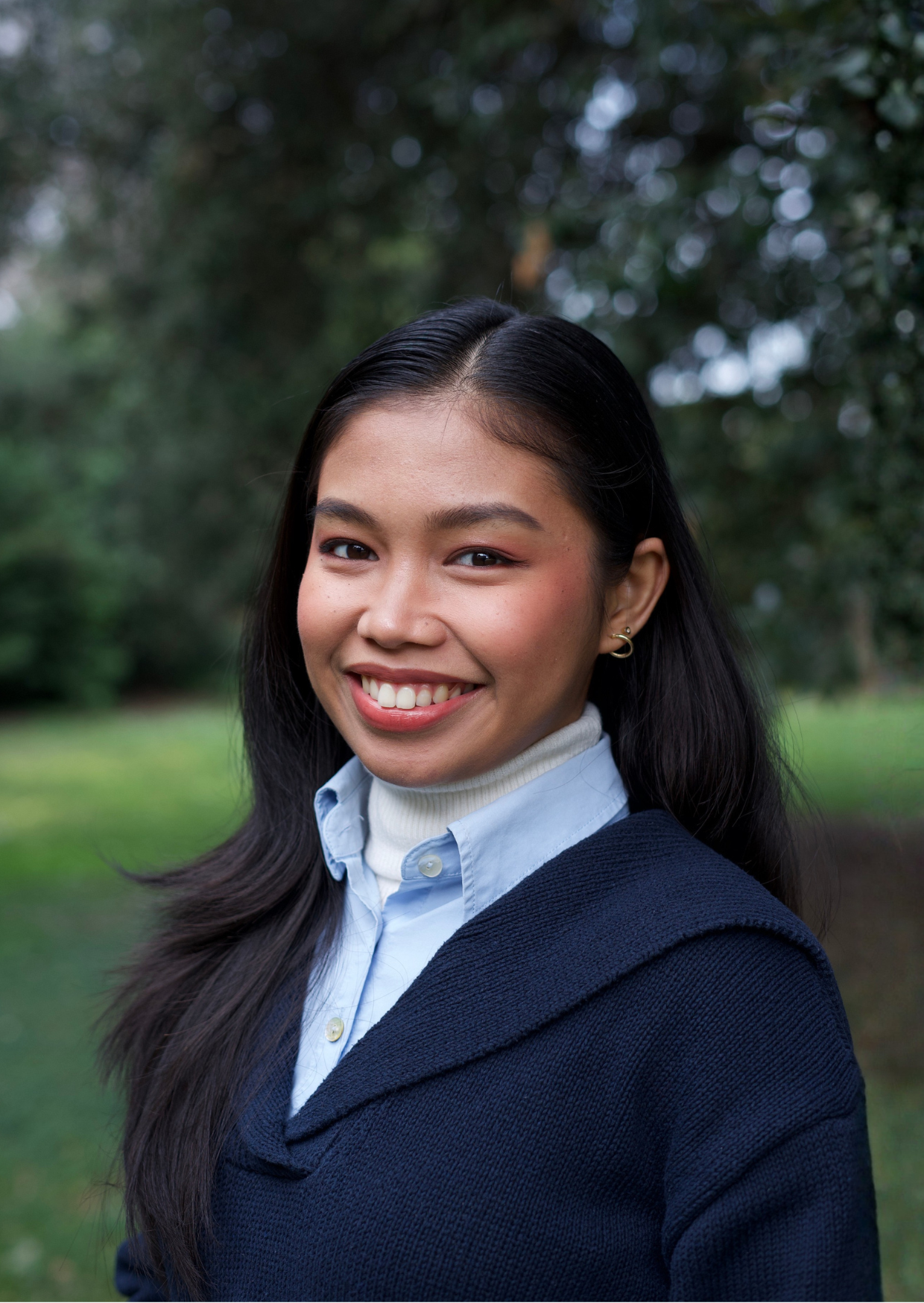Young person with long dark hair in a navy sweater over a blue collared shirt, outdoors in a park.