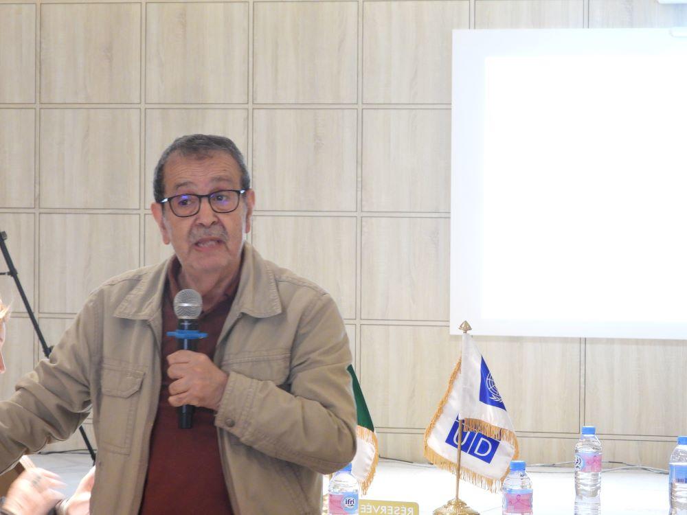 Man speaks into a microphone in a conference room; projector screen and Lions banner visible.