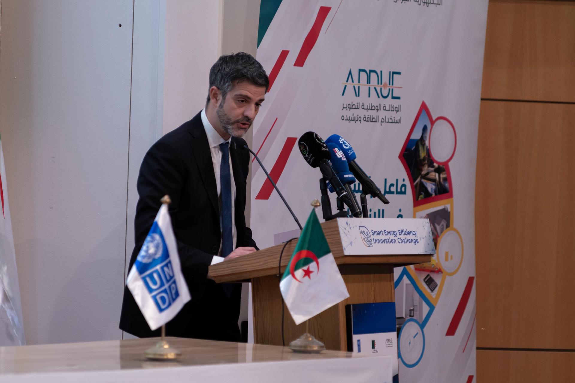 Speaker in suit at podium with microphones, banners, and flags in a conference room.