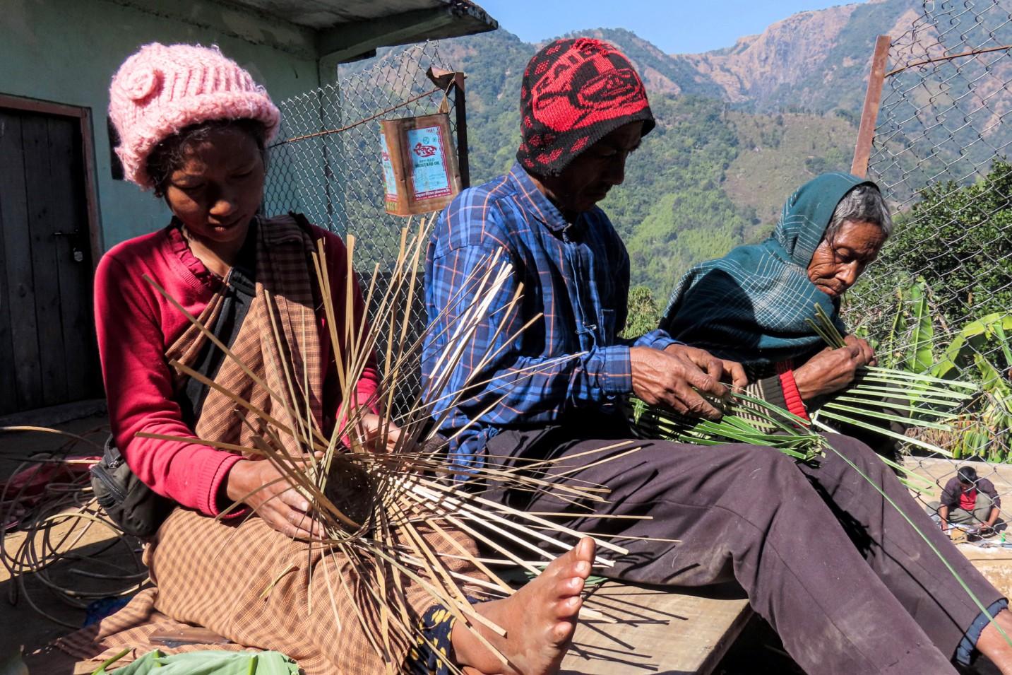 Three people weaving dried grass outdoors on a hillside with mountains in the background.