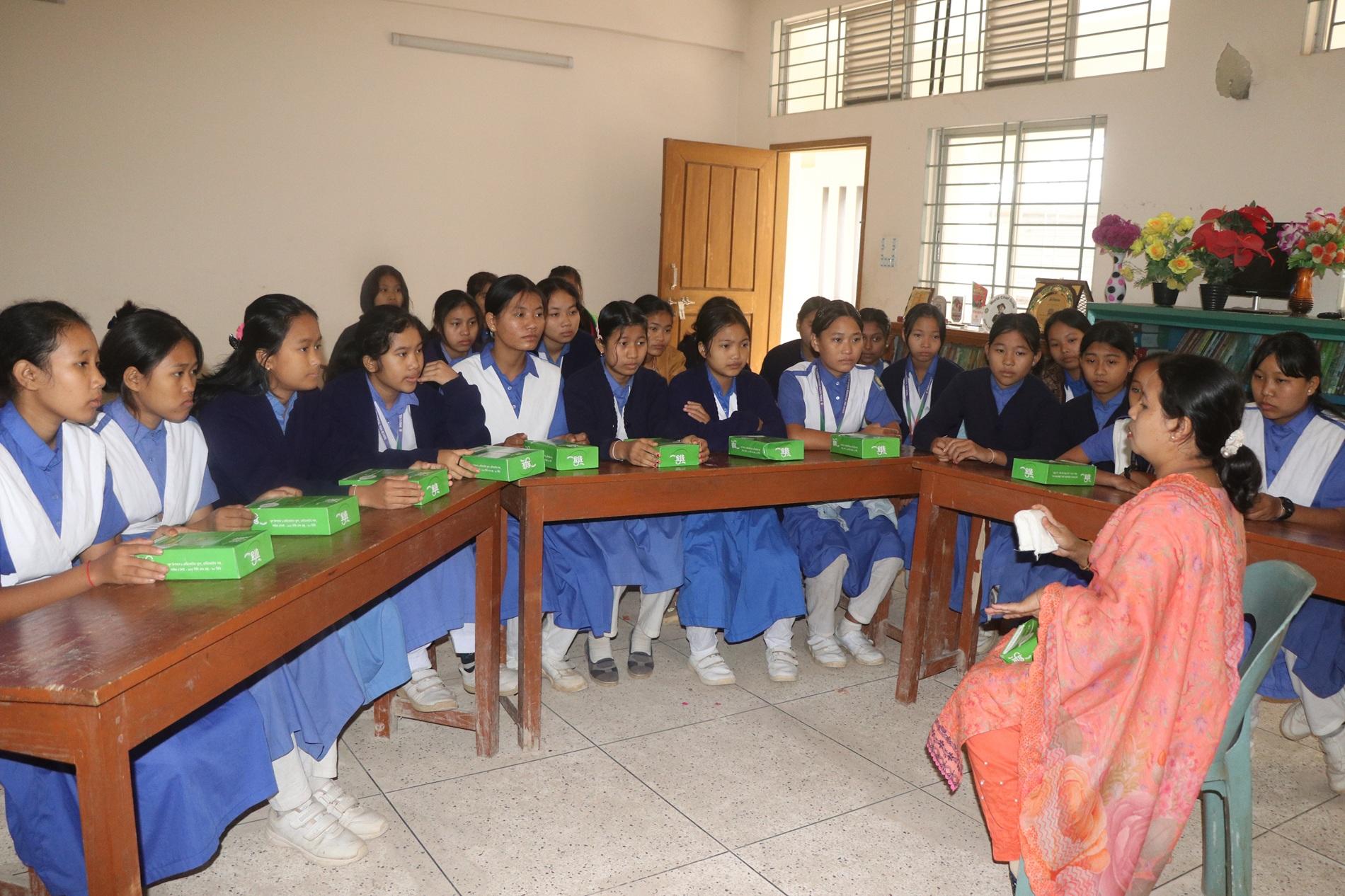 Female students in blue uniforms sit around desks in a classroom; a woman in pink leads at front.
