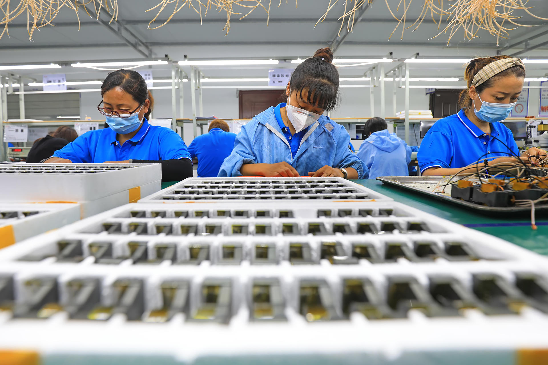 Photo: workers in blue uniforms and masks assemble electronics on a production line.