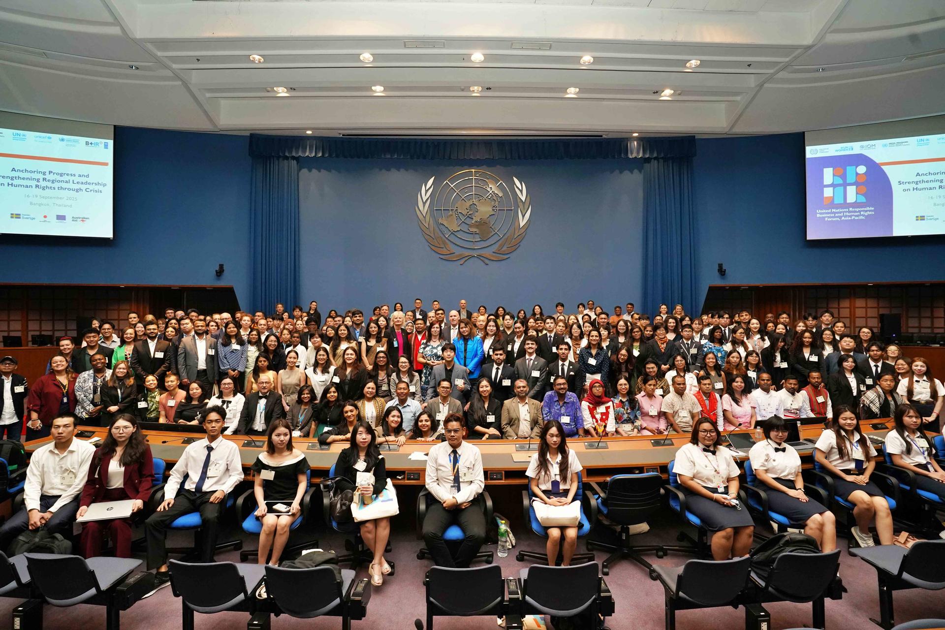 Crowded assembly hall with delegates wearing badges, UN emblem on a blue backdrop.