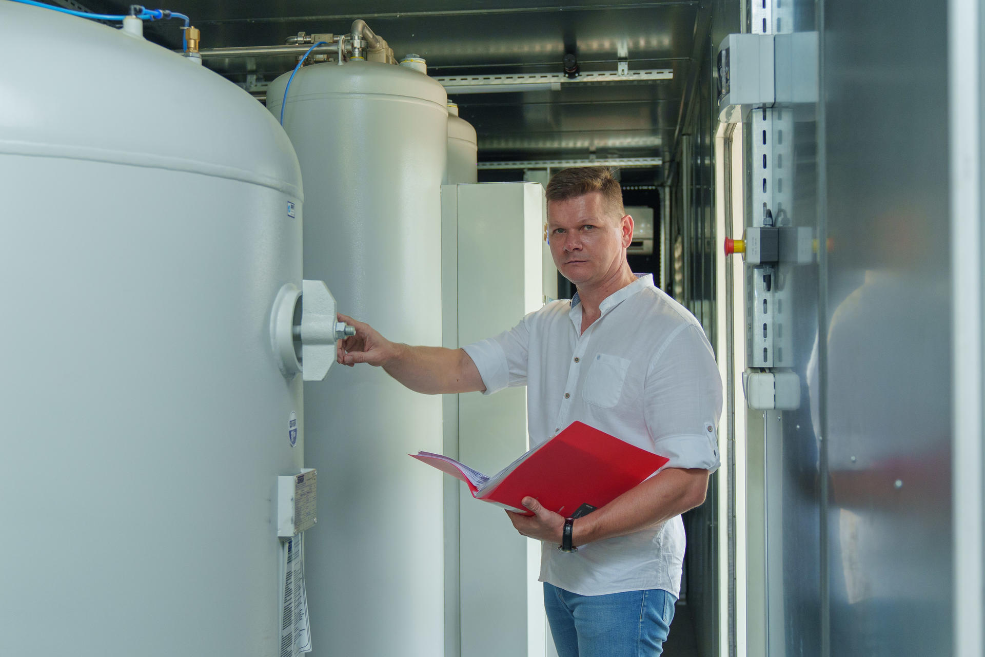 Man in a white shirt holds a red folder while inspecting large industrial tanks.