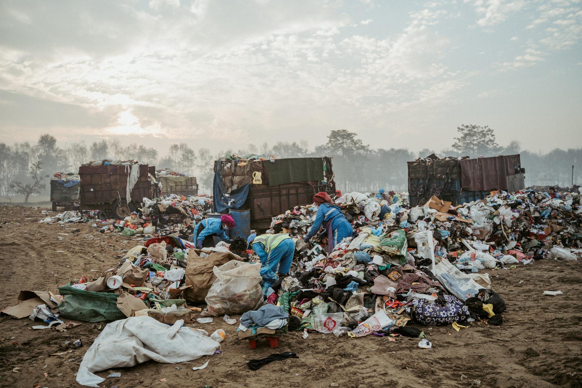 People sorting trash at a large landfill with piles of garbage under a cloudy sky.