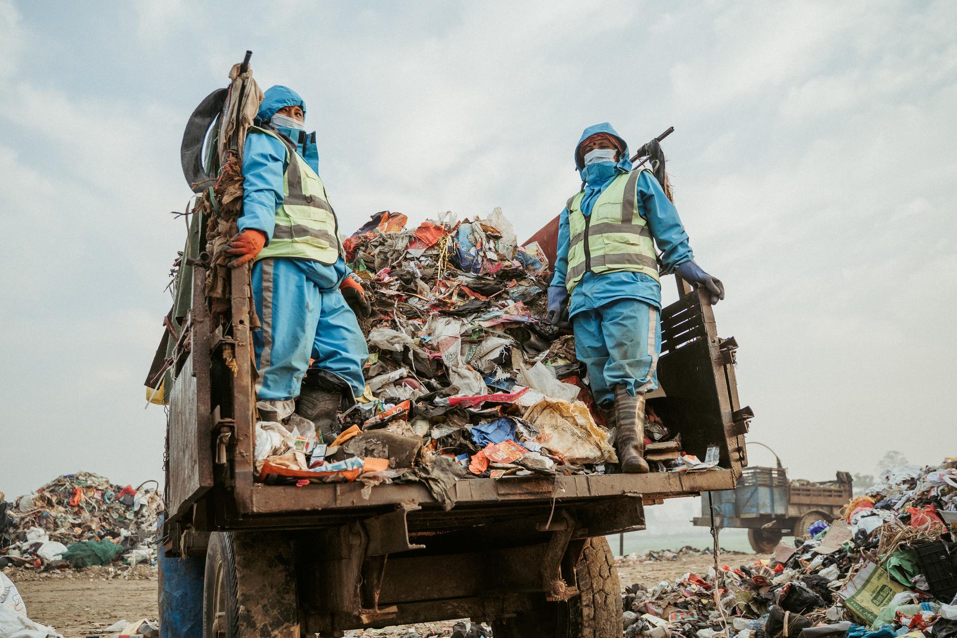Two workers in blue uniforms stand on a dump truck loaded with trash at a landfill.