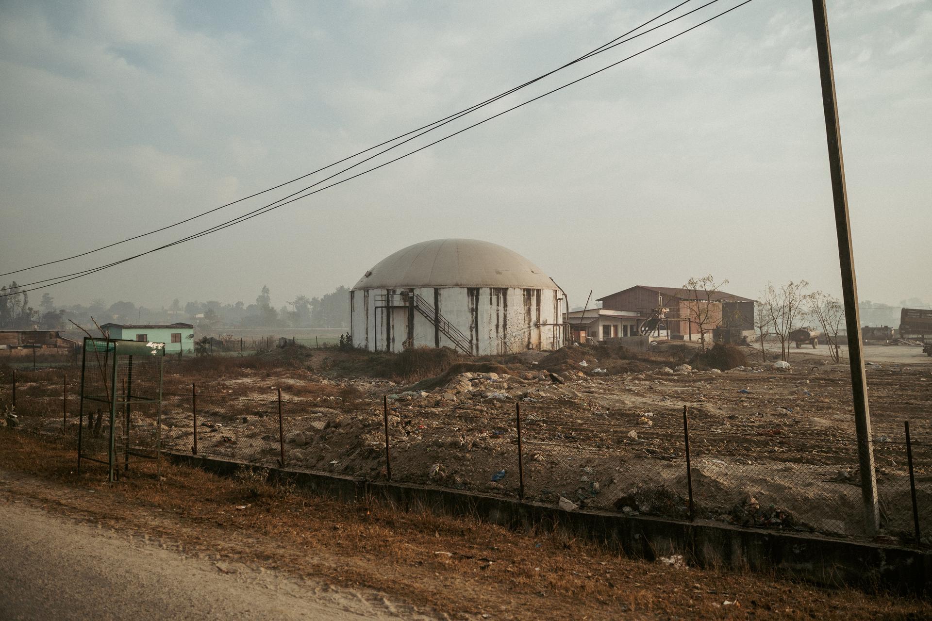 Dusty landscape with a white domed building behind a fence, power lines, and scattered houses.