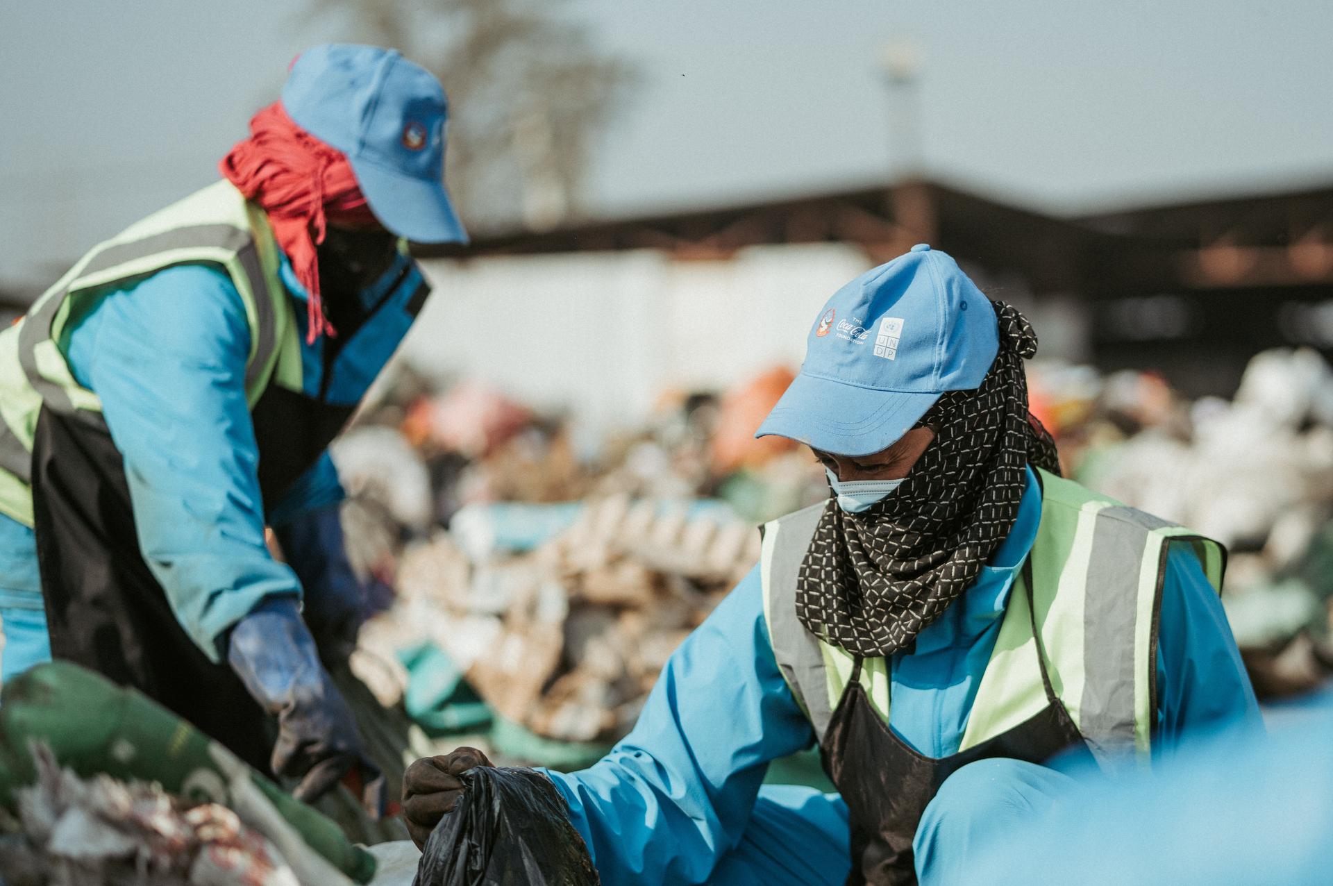 Two workers in blue uniforms and reflective vests shake hands outdoors, crowd in background.