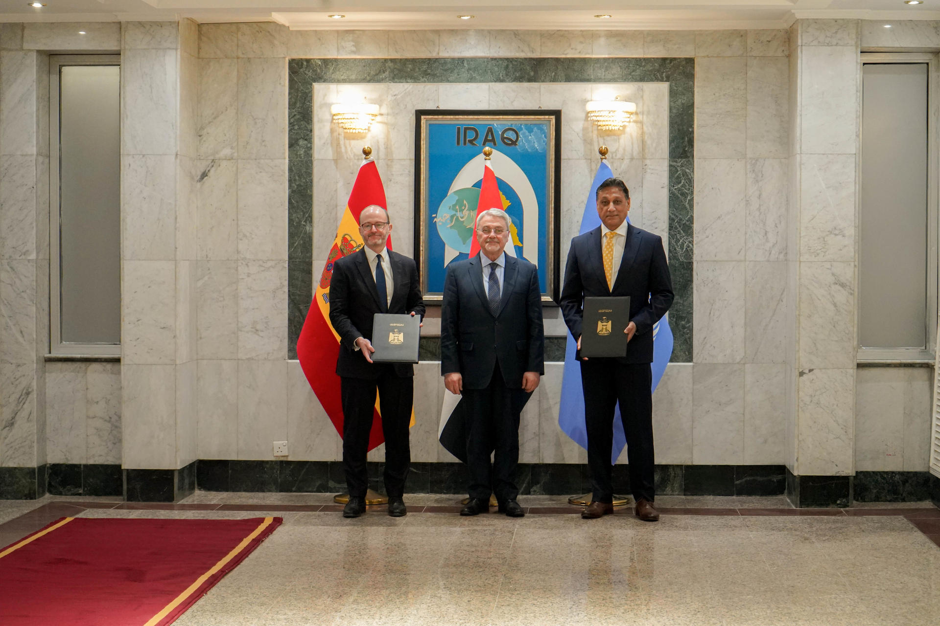 Photograph of three formally dressed officials in a marble hall, holding folders, with a crest and flags behind.