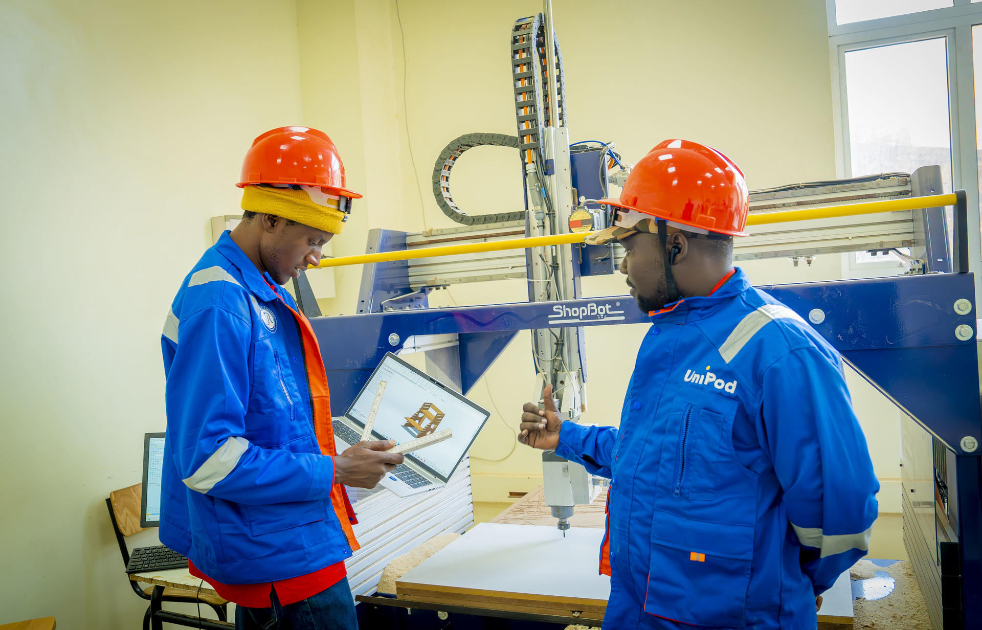 Two workers in blue coveralls and orange hard hats inspect equipment in a workshop.