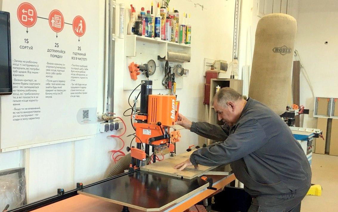 Photograph of a man in a workshop leaning on a workbench beside an orange machine.