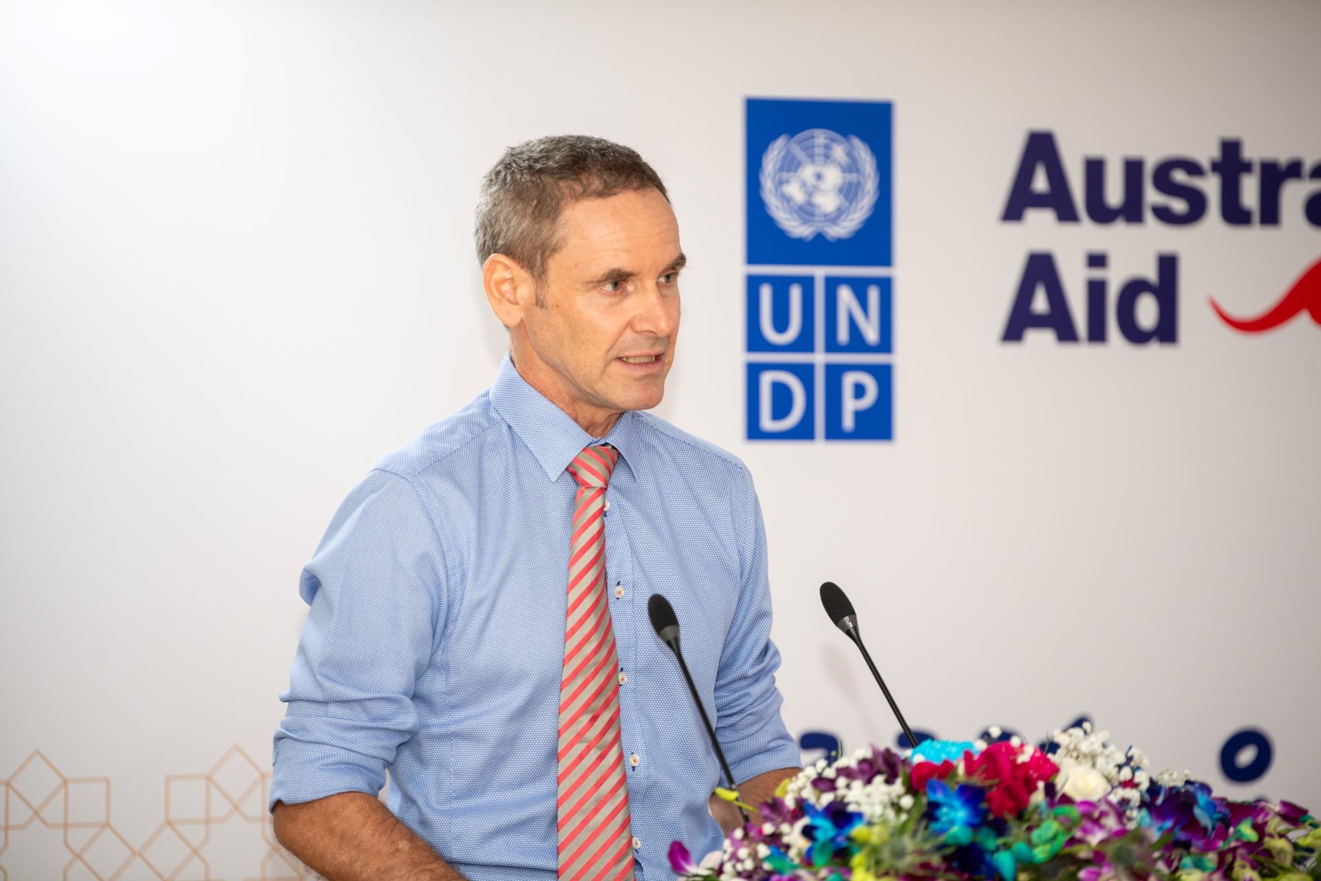 Speaker in blue shirt and pink striped tie at podium with UNDP and Australian aid banners, flowers.