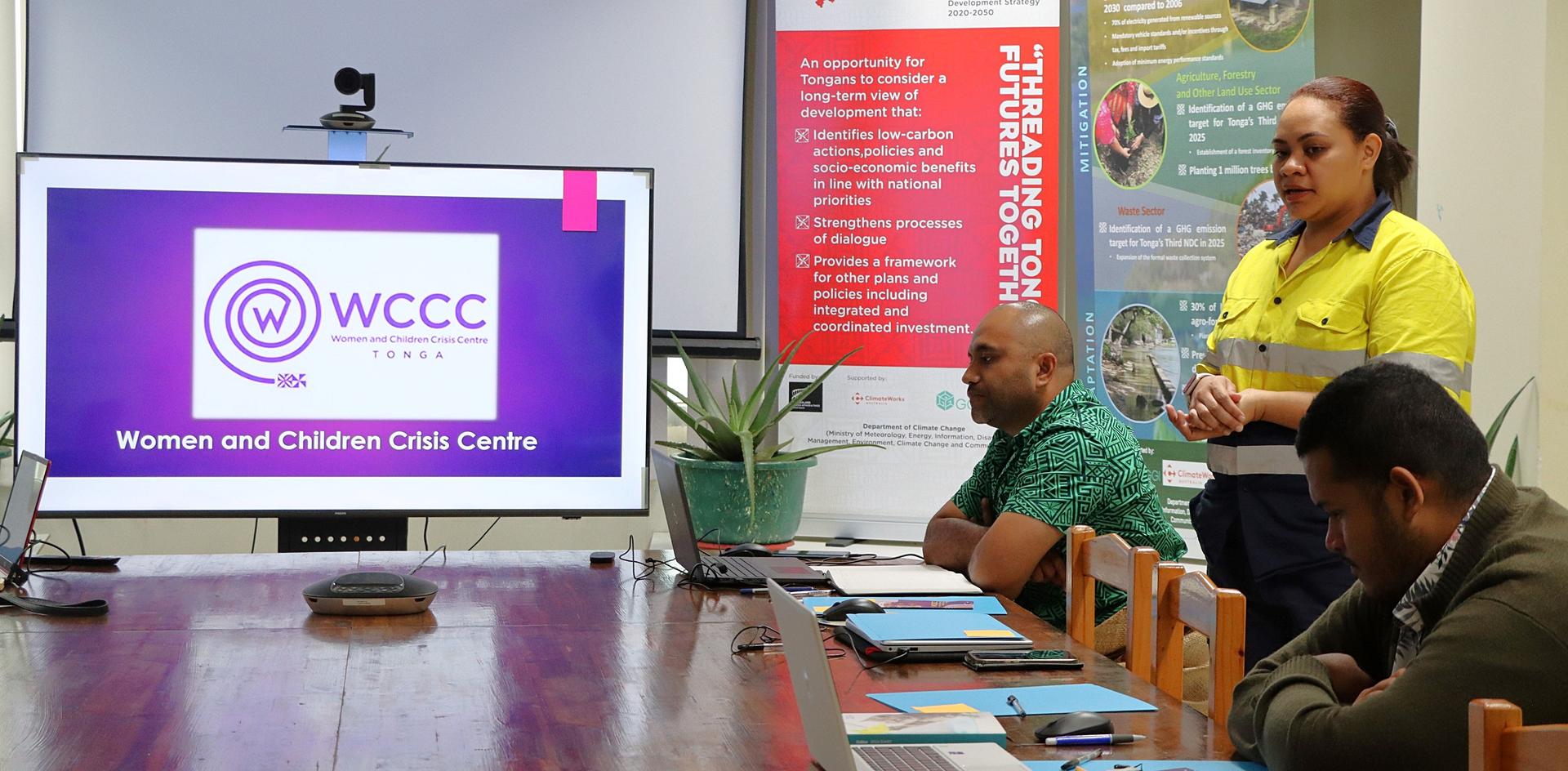 Conference room with a presenter in yellow beside a screen showing a WCCC logo; attendees sit at a long table.