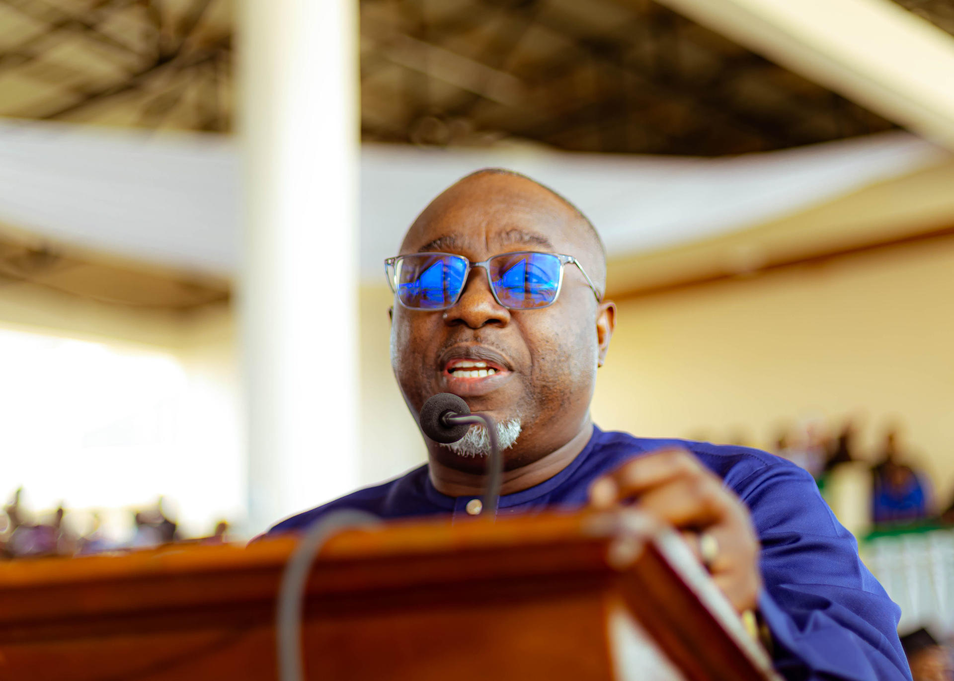 Man speaks at wooden podium in blue shirt, audience in background.