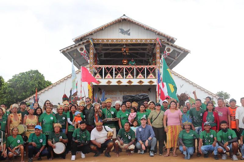 Group of people on an outdoor stage in front of a wooden pavilion, many wearing green shirts.