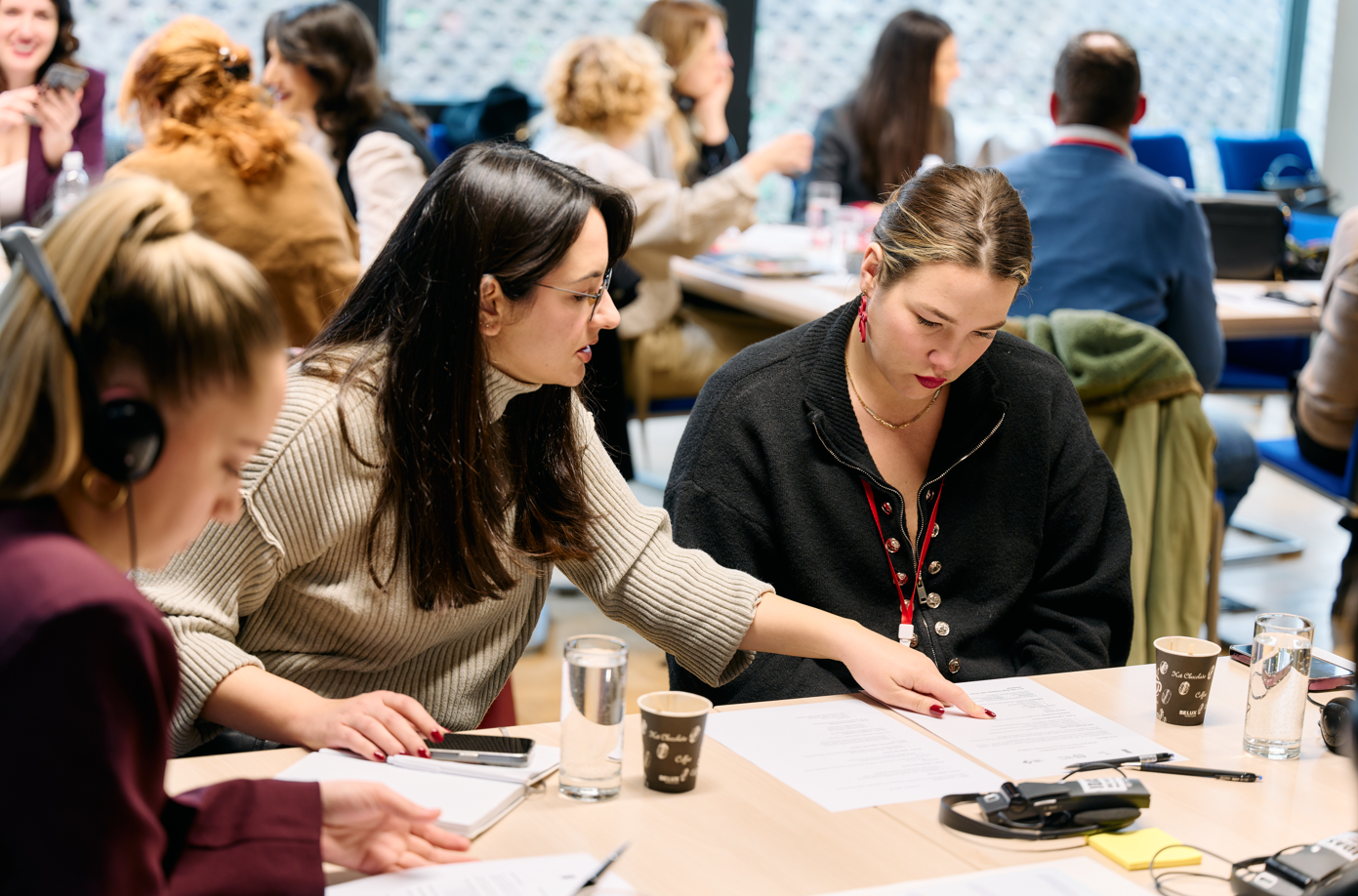 Group of students at a classroom table, writing and discussing; coffee cups and notebooks.