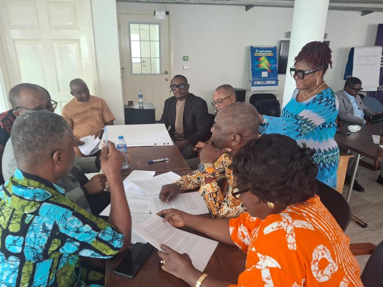 Photo shows a group of people in colorful outfits around a conference table, discussing papers.
