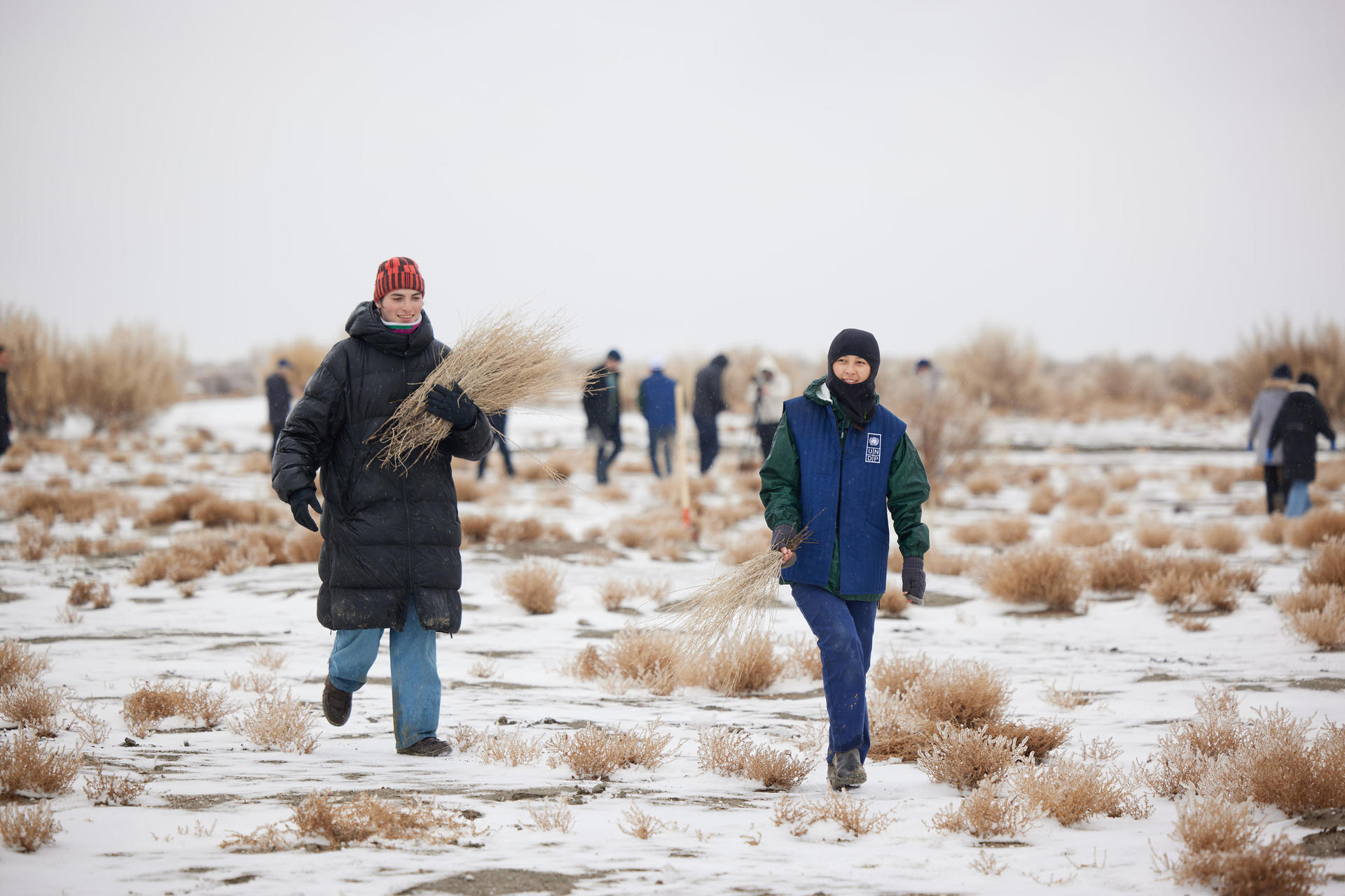 Photograph of a group of people in winter coats walking across a snow-dusted field with dry grass.