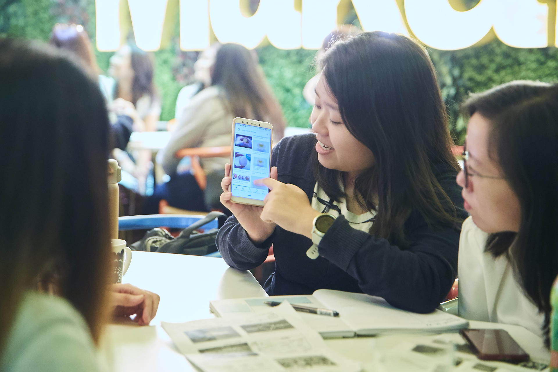 Group of people at a table examining papers; central person shows blue smartphone screen.