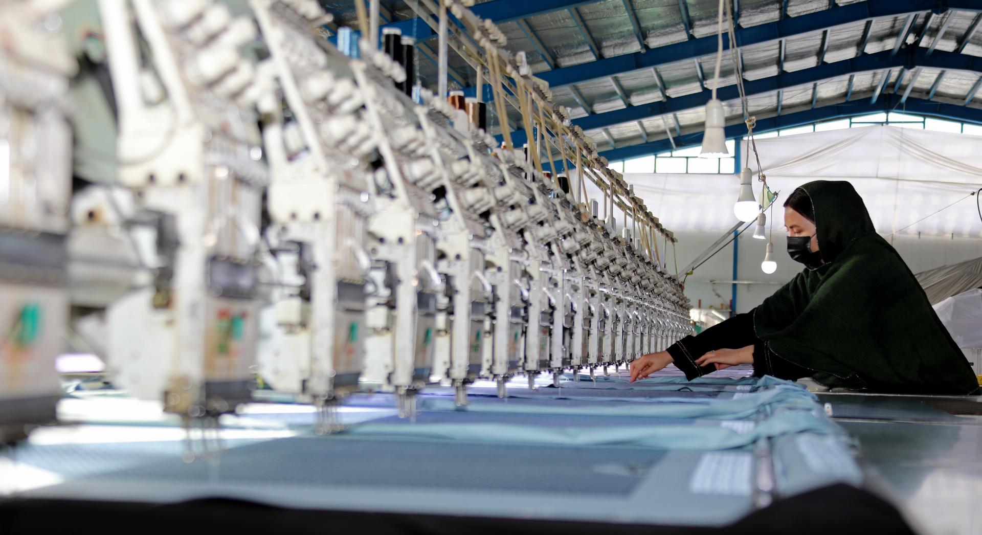 Worker in a green hijab sits at a long row of sewing machines in a textile factory.