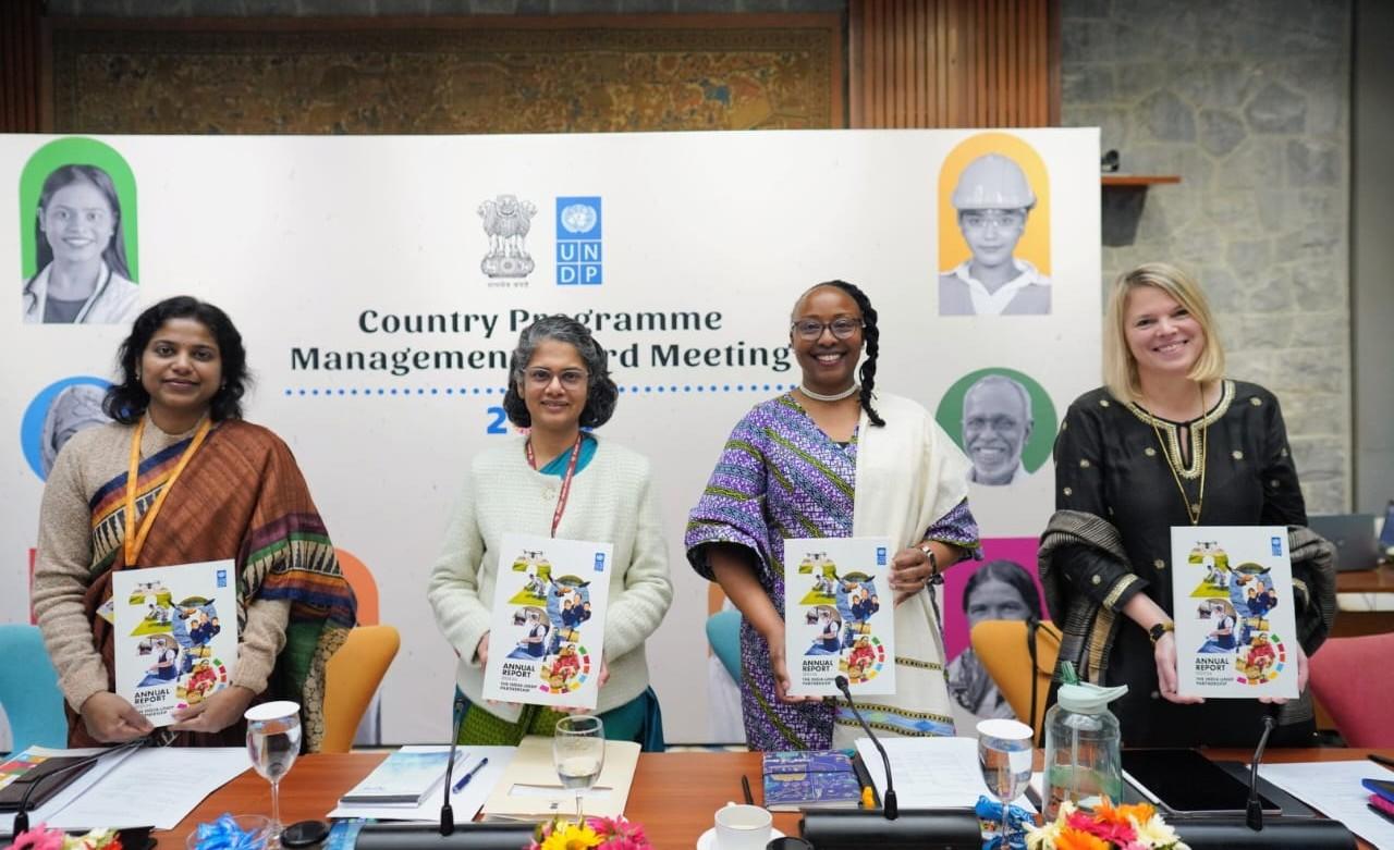 Five women in colorful outfits stand behind a conference table with documents and water, a banner in the background.