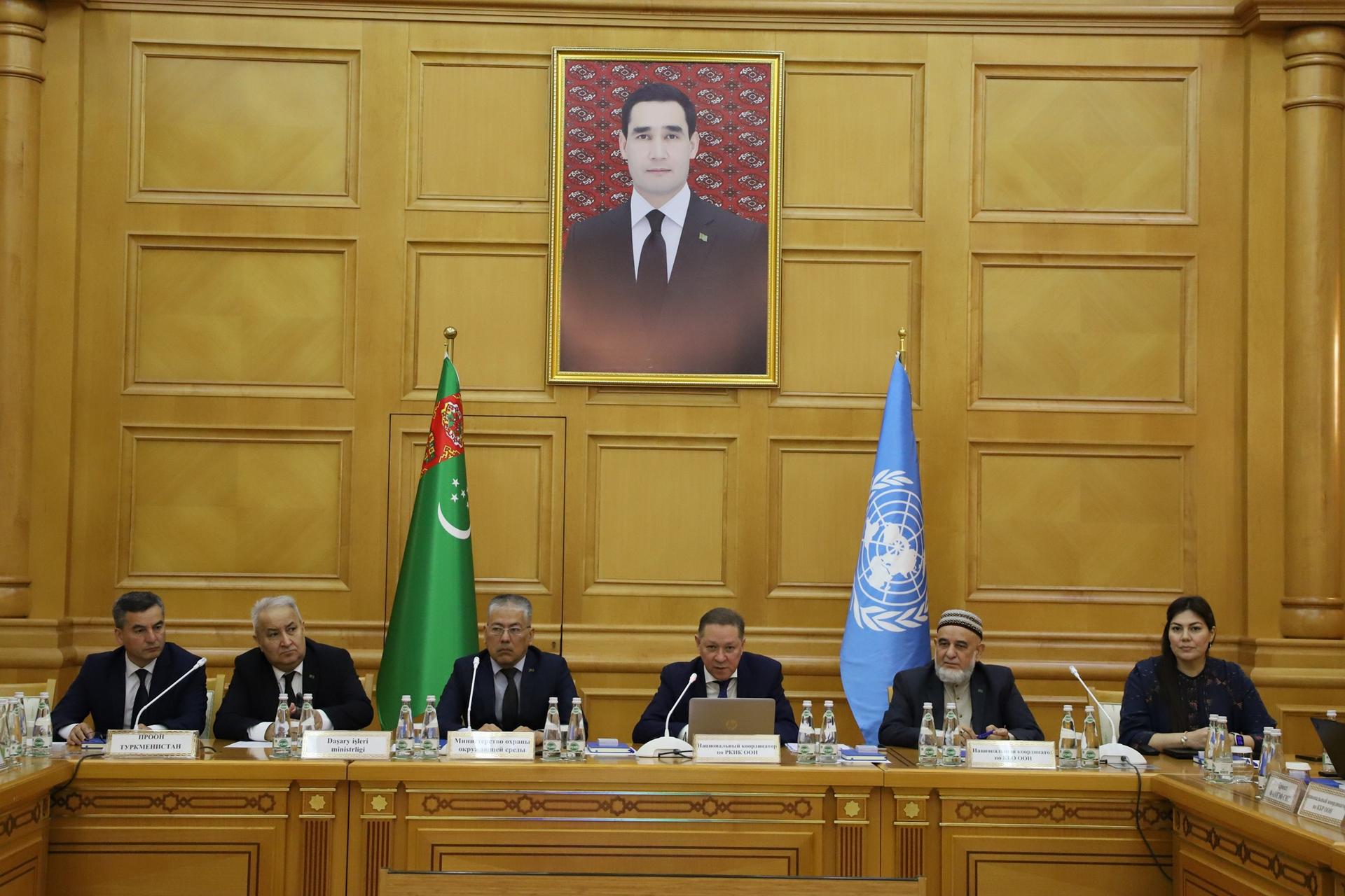 Officials seated at a long wooden table in a wood-paneled room with two flags and a portrait.