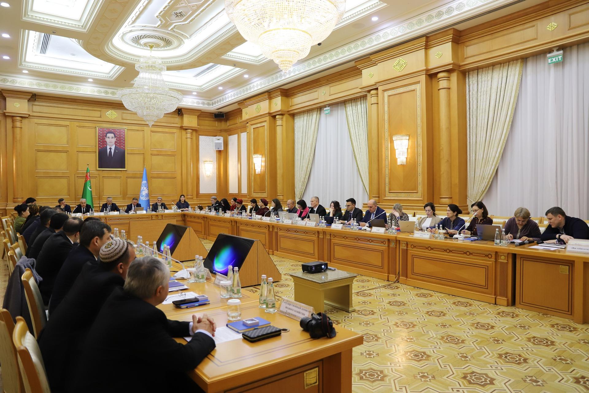 Formal conference room with a long U-shaped wooden table, numerous attendees, chandeliers, and gold curtains.