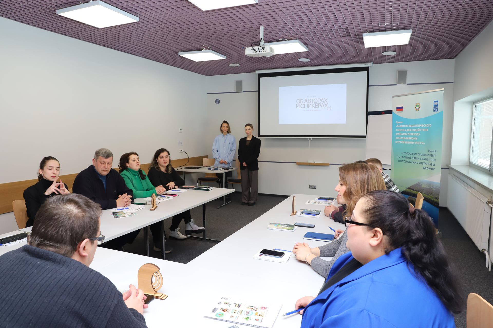 Group of people around a U-shaped table in a classroom; presenter at the front with a projector.
