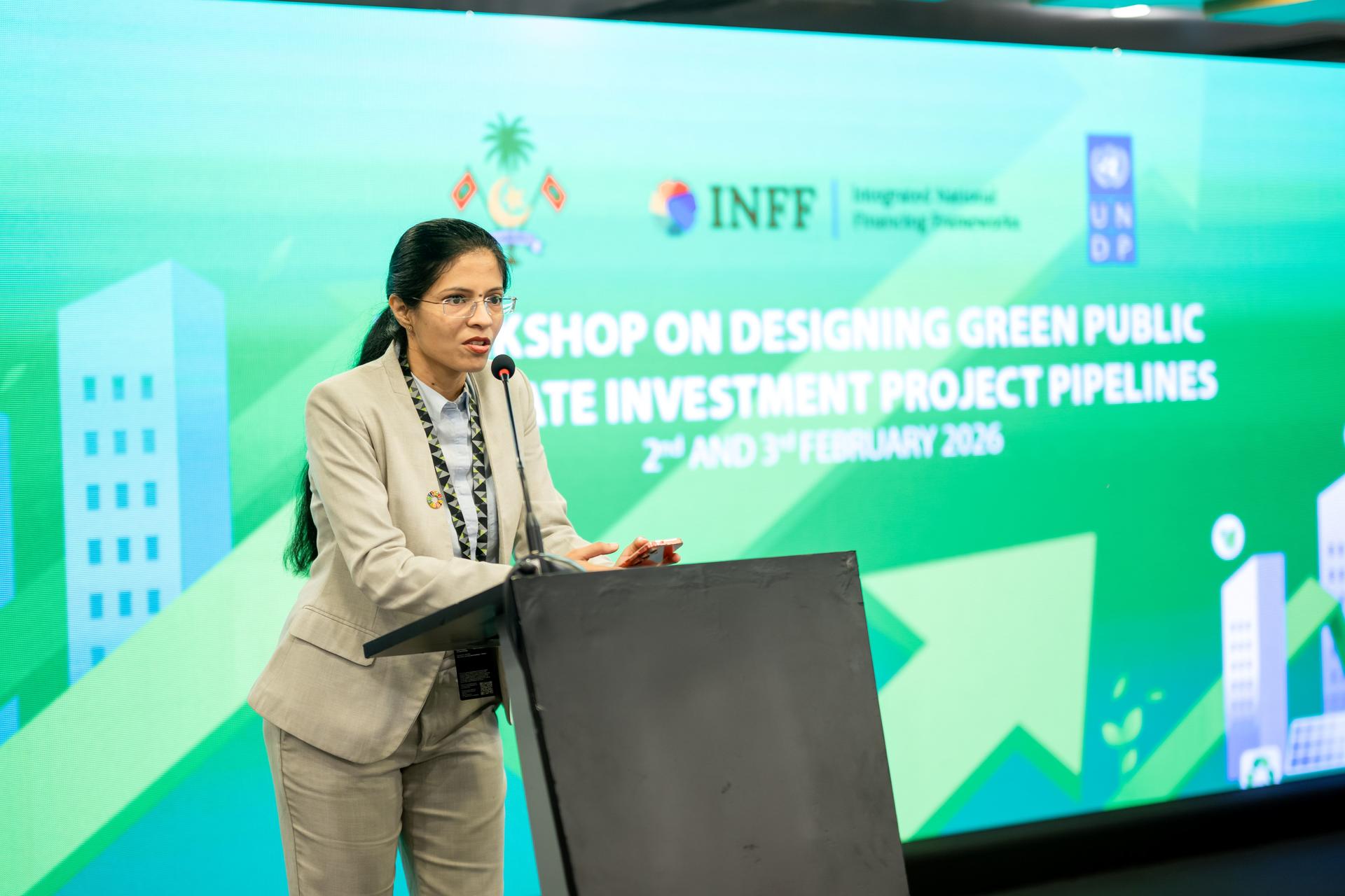 Woman in gray suit speaks at a podium on a stage with a green-blue geometric backdrop.