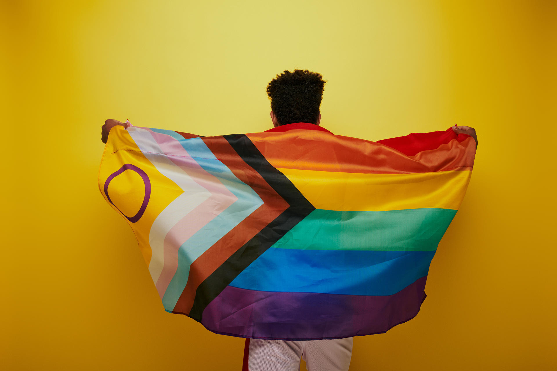 Photograph of a person with back to camera holding a rainbow flag against a yellow background.