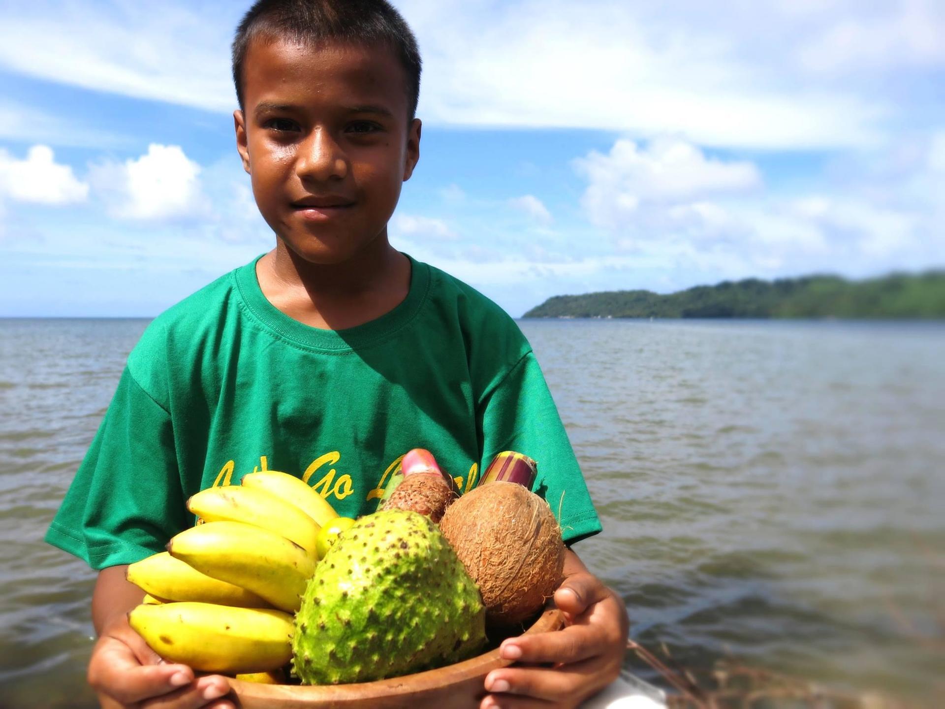 Man in a green shirt on a beach holds bananas and a durian with the sea in the background.