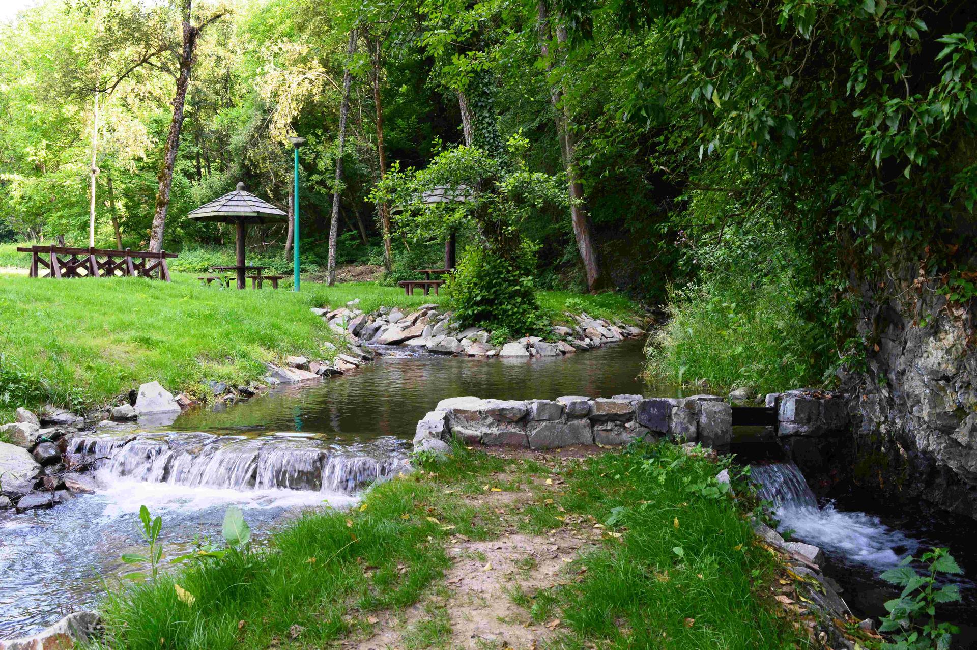 Woodland scene with a clear stream, rocks, and a small wooden shelter in the background.