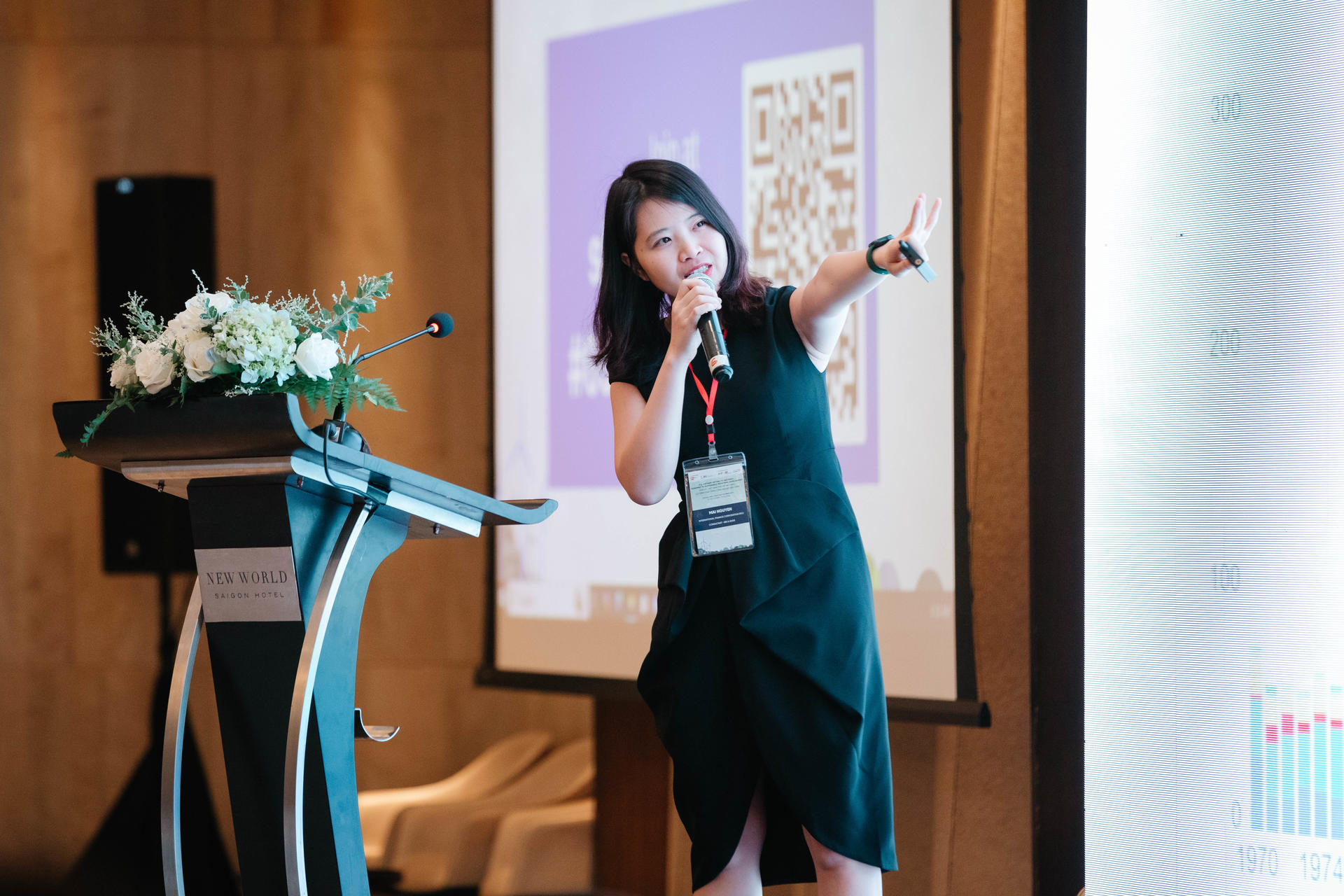 Female presenter on stage gesturing with microphone; podium with flowers and a slide screen behind.