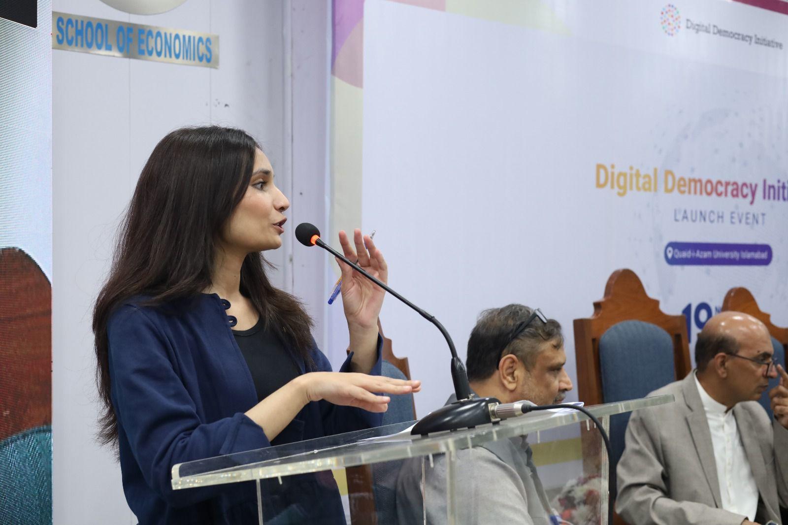 Speaker at podium in blue blazer; panelists sit on stage with Digital Democracy banner.