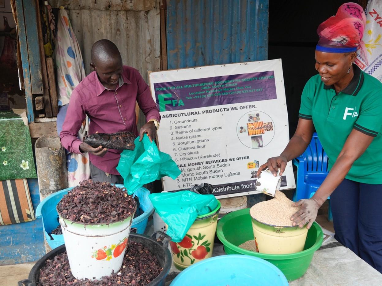 Two market vendors with baskets and sacks of produce beside a signboard.