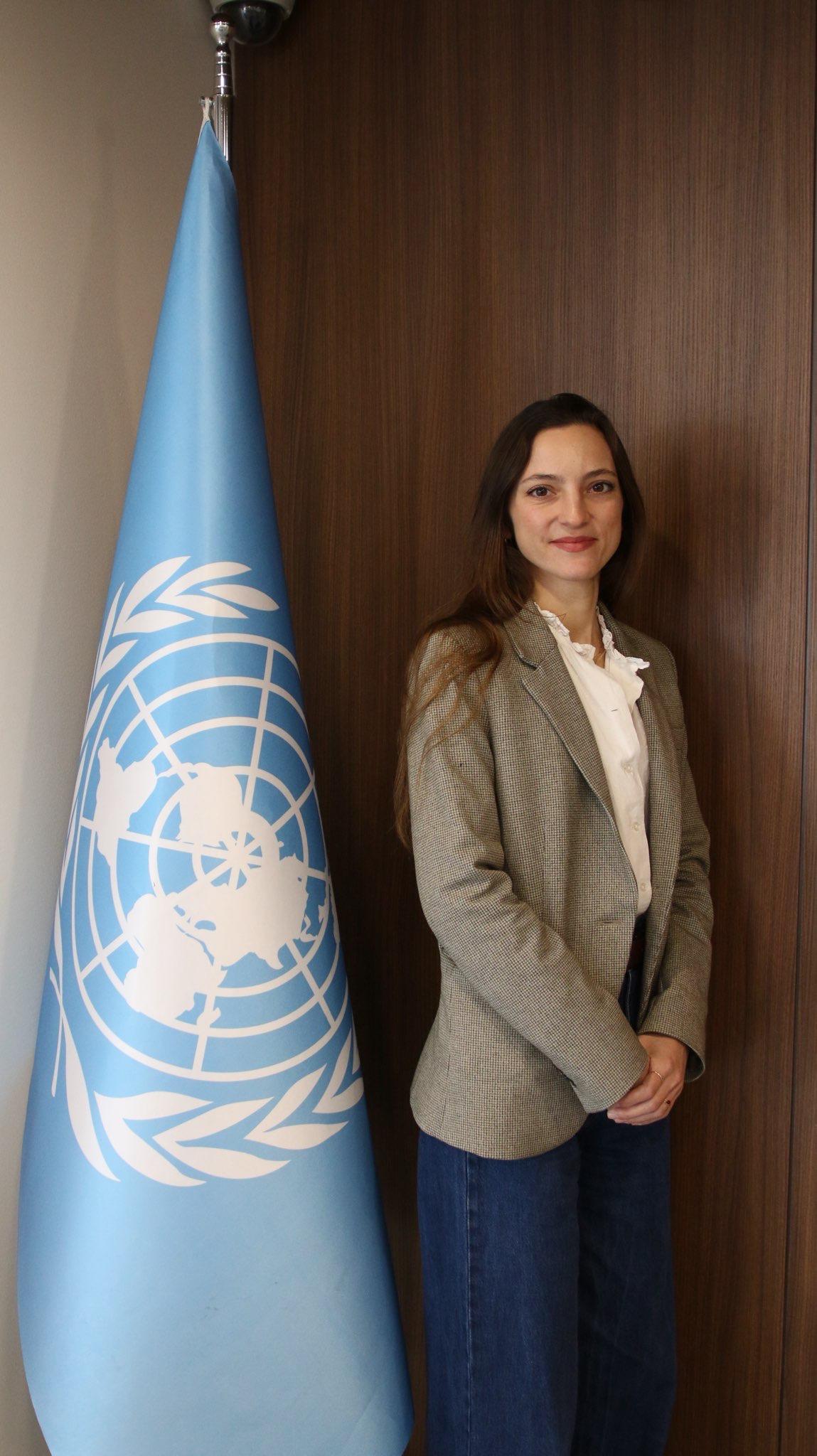 Woman in a beige blazer standing beside the United Nations flag against a wood-paneled wall.