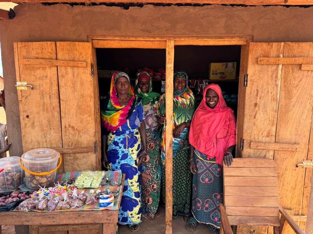 Three women in bright sarees stand in a mud-brick doorway beside a market stall.