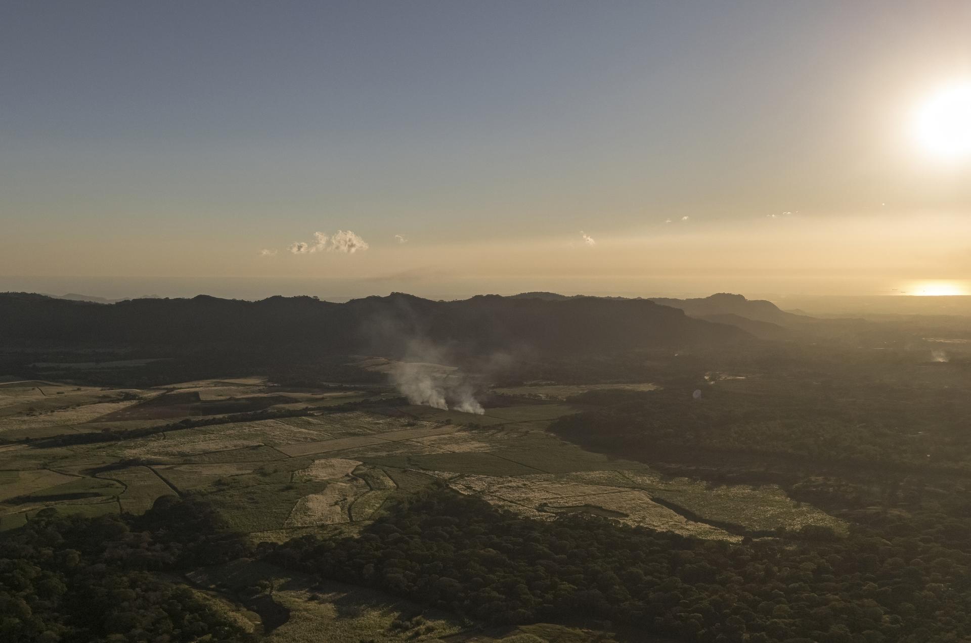 Sunset landscape over fields and distant hills with a small smoke plume.